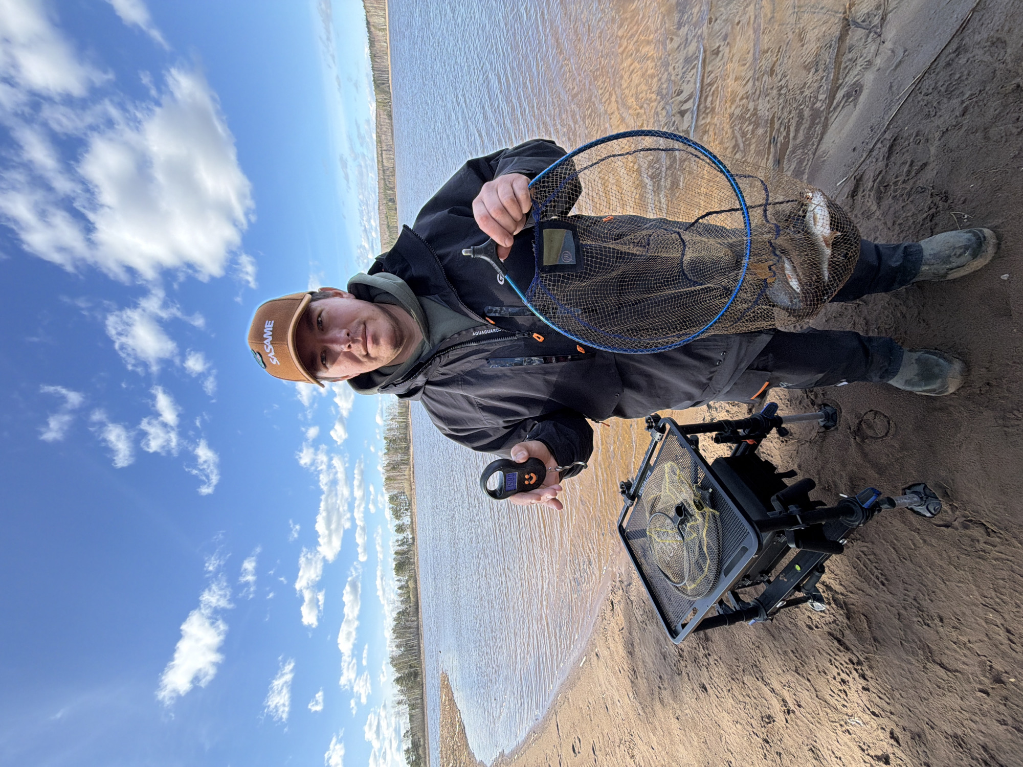 A man stands on a sandy bank alongside a body of water, likely a river or lake, under a bright blue sky with scattered clouds. He wears a brown cap, dark jacket, and black pants tucked into dark green boots. He is holding a fishing net containing several fish and a digital scale in his other hand, displaying the weight of the catch. A black fishing platform with a net rests on the sand near him. The shoreline extends into the distance, and a line of trees is visible on the opposite bank. The location is likely in a natural area near the city of Chaikovsky, Russia.