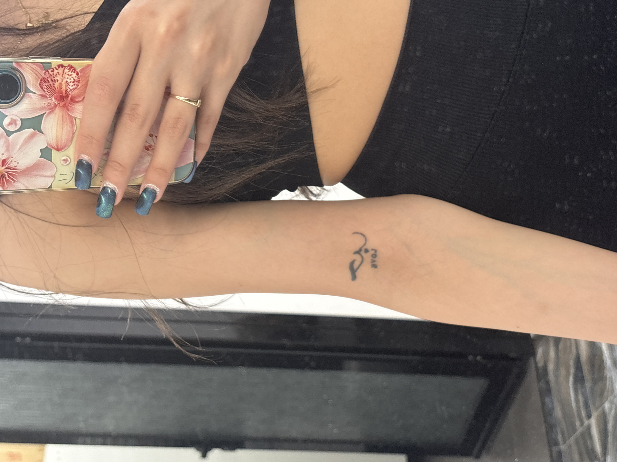 A close-up shot focuses on a person's left arm, displaying a small tattoo on the inner forearm. The tattoo appears to be a minimalist design, featuring abstract shapes alongside the word "love" written vertically. A section of the person's black top is visible in the upper right corner of the frame. The person is holding a phone with a decorative case featuring pink floral designs. The hand holding the phone is adorned with a gold ring and blue glittery nails. The background consists of a blurred surface, reflecting light and obscuring details. The location and city are not determinable from the image.