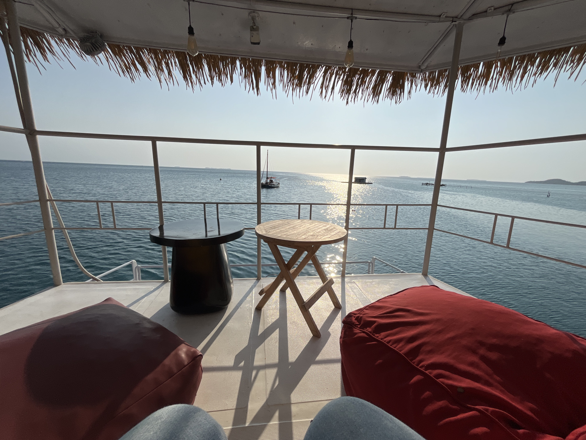 The image depicts a serene, open-air seating area on what appears to be a boat or a floating platform, overlooking a calm blue ocean under a clear sky. In the foreground, two large, plush red beanbag chairs are positioned on a white deck. Between them, a small, foldable wooden table stands, casting a geometric shadow on the deck. To the left of the table, a dark, cylindrical table with a glossy black top is present.
The deck is bordered by a white railing. Beyond the railing, the ocean stretches out to the horizon, reflecting the sunlight. In the distance, a small sailboat is visible, along with a floating structure and other distant boats or buoys. The overhead structure of the seating area is covered with dried palm fronds, providing a natural shade. Two exposed light bulbs hang from the ceiling.
The scene is bathed in soft, natural light, suggesting it might be morning or late afternoon. The overall impression is one of relaxation and tranquility, with a clear view of the seascape.
This scene is located in Karimunjawa, Indonesia. Karimunjawa is an archipelago of 27 islands located in the Java Sea, north of the city of Jepara on Java Island. It is known for its pristine beaches, clear waters, and vibrant coral reefs, making it a popular destination for diving, snorkeling, and relaxation. The open-air seating area with a view of the ocean is a common feature in resorts and accommodations found in this tropical paradise.