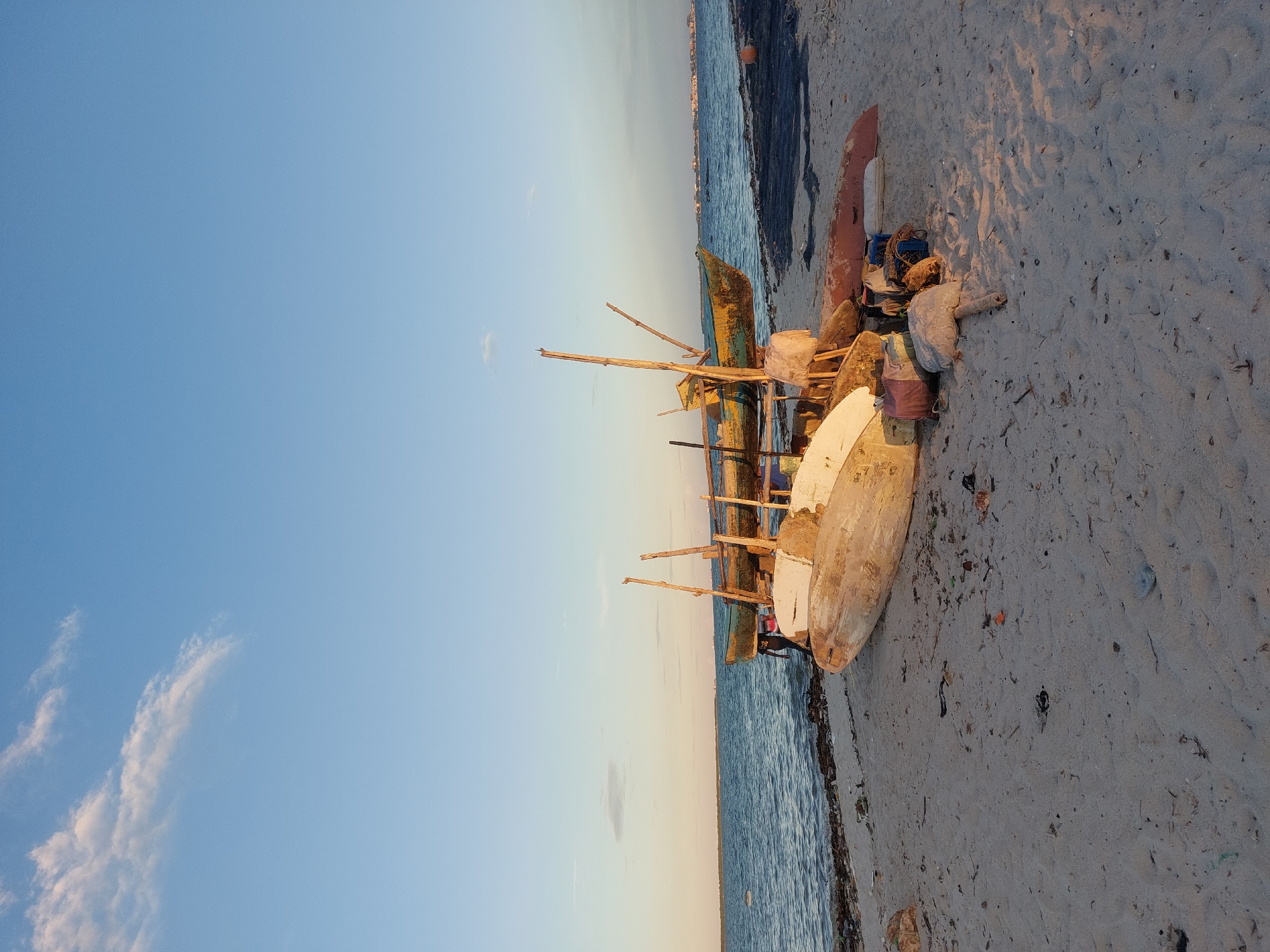 The image depicts a beach scene in Dar es-Salaam, Tanzania. Several wooden fishing boats are resting on the sand near the shoreline. The boats are of varying sizes and appear weathered. Beach debris is scattered around the boats. The sky is partly clear with a light blue gradient. The water is a darker blue. The sand is a light gray color. There are no people visible in the image.