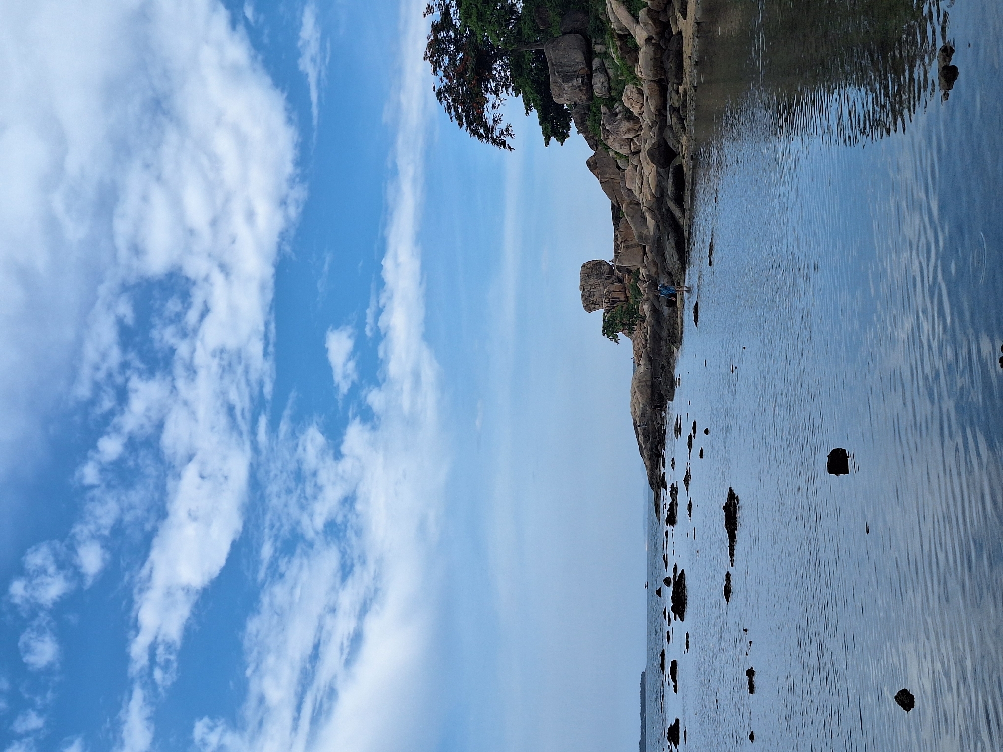 The image depicts a coastal scene, likely in Nha Trang, Vietnam, characterized by a rocky shoreline bordering a tranquil body of water. The water's surface is rippled, reflecting the blue sky and scattered white clouds. Numerous dark rocks of varying sizes are present in the water and along the shore.
On the rocky embankment, there is a person wearing blue shorts and a light-colored shirt, standing in shallow water near the edge. Further inland, a distinctive large boulder rests atop a cluster of other rocks, with lush green trees and a tree with red flowers growing on the higher ground. In the distance, across the water, a small island or landmass is visible, with a boat or ship in between. The sky is a vibrant blue, with wispy white clouds creating a dynamic overhead backdrop. The overall impression is of a serene, natural landscape under a bright sky.