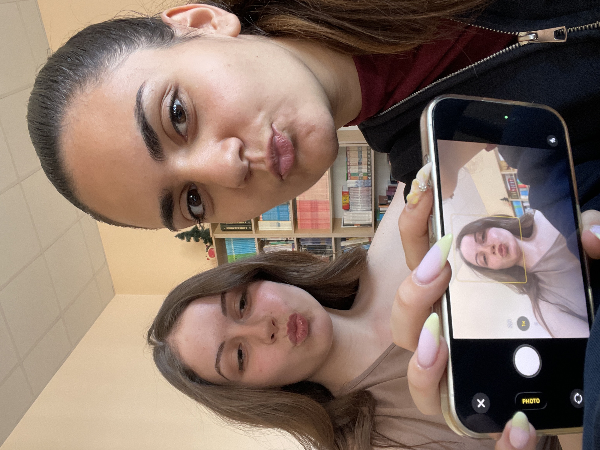 Two women are taking a selfie indoors. The woman at the top has dark hair pulled back, wearing a black hoodie over a dark red top, and making a kissing face. The woman at the bottom is holding a smartphone with a gold case and long nails. The smartphone screen shows a live image of the woman taking the picture, also making a kissing face. A bookshelf filled with books is visible in the background. The location is indoors and possibly in Burgas, Bulgaria, though there are no elements in the picture to strongly confirm this.