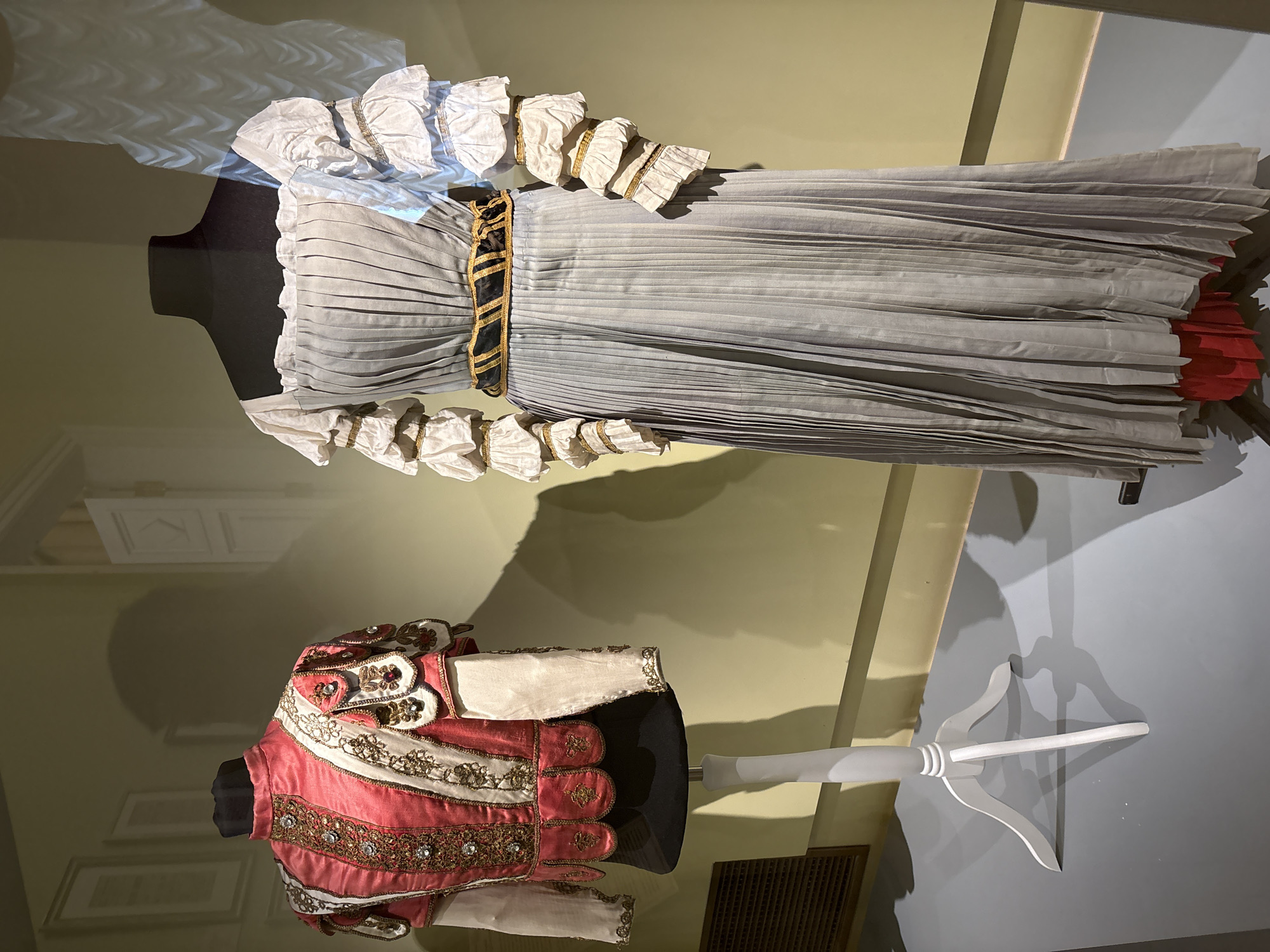 The image shows two garments displayed in a museum setting, potentially in Saint Petersburg, Russia, although this cannot be confirmed from the image alone.
The top garment is a long, pleated dress, predominantly light grey/blue. It features elaborate white ruffled sleeves and neckline trim with a gold accent. A dark belt with gold trim cinches the waist. The dress is displayed on a black mannequin.
Below the dress is a shorter, ornate jacket, primarily red with white and gold embellishments. It also features intricate detailing and is displayed on a black mannequin with a white stand.
The background consists of a light green wall with a hint of a doorway in the upper left corner. The bottom right corner shows a light grey/blue wall and a white stand.