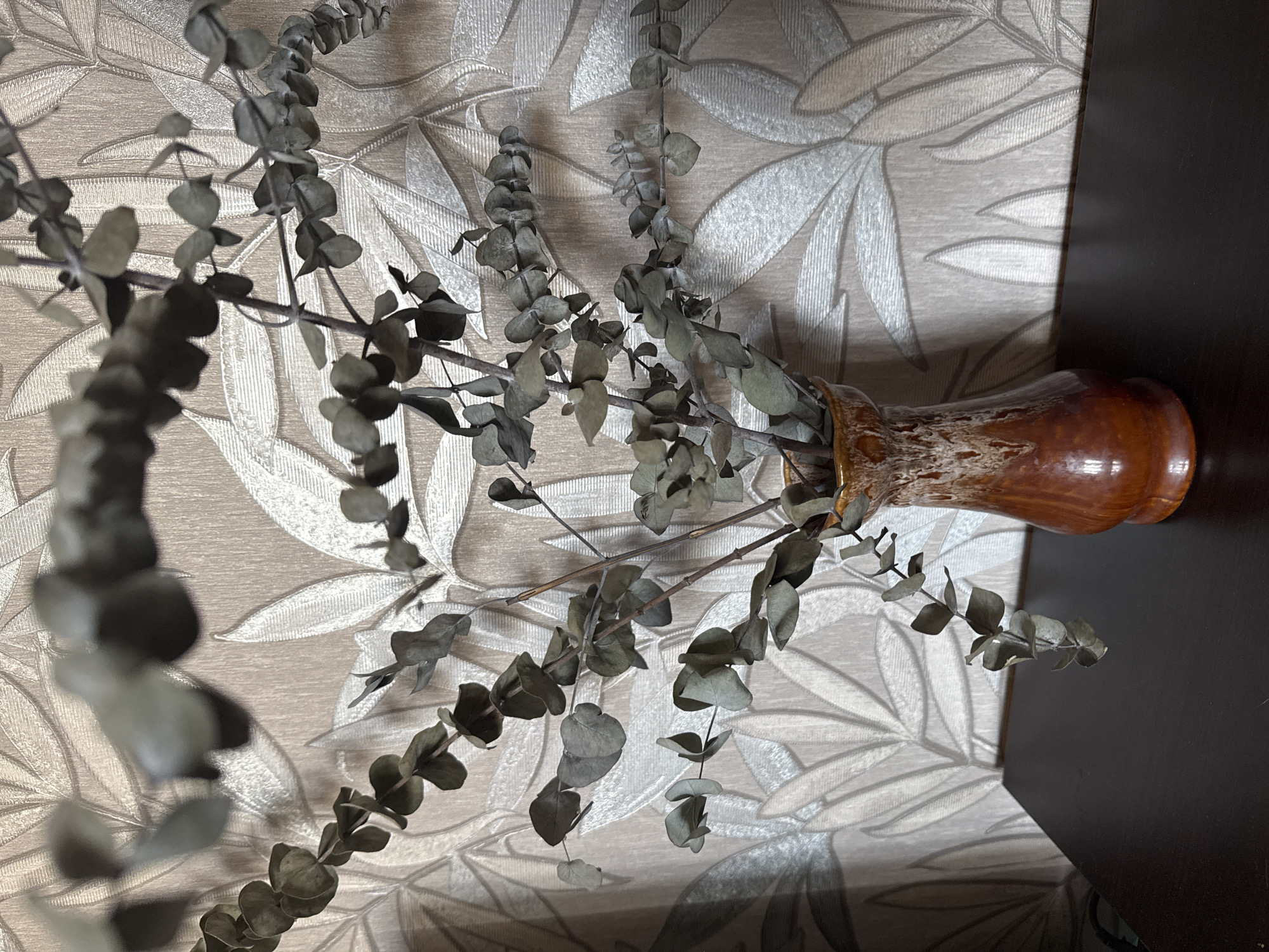 A close-up shot captures a vase containing dried eucalyptus branches, set against a beige, textured wallpaper with a leaf pattern. The vase, made of glazed ceramic, displays a gradient of brown and cream colors. The dried eucalyptus branches, with their small, round, grey-green leaves, extend upwards and outwards from the vase, some reaching towards the top of the frame. The lighting casts shadows on the wallpaper, highlighting the embossed leaf design. The foreground is dominated by a dark brown surface, likely a table or shelf, on which the vase rests. There are no people or animals present in the scene. The image appears to be taken indoors, possibly in a domestic setting, within Bishkek City, Kyrgyzstan.