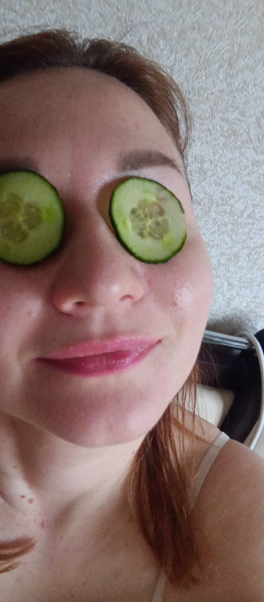 A woman is shown in a close-up, face-level view. Her eyes are covered with slices of cucumber. Her hair is reddish-brown. She is wearing lip gloss and her skin has visible freckles. The background consists of a wall with a textured surface and a blurred object that appears to be a chair or headrest. The city is Unknown, Unknown.