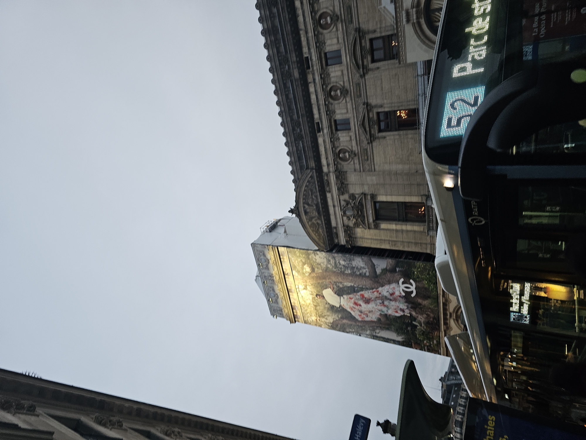 The image depicts a street scene in Paris, France, specifically at the intersection of Rue Halévy, identifiable by the visible street sign, and adjacent to the Opéra Garnier, which is recognizable by its distinct ornate facade on the right.
The scene is viewed from a low angle, looking upward towards the buildings and an overcast, light grey sky that occupies the upper third of the frame. In the foreground, the upper front portion of a white and grey RATP public transit bus is visible. Its digital display shows the illuminated number "52" on the left and "Parc de st" on the right, indicating its route and destination. The bus windshield and side windows reflect ambient light, and interior illumination is discernible.
To the left of the Opéra Garnier stands a multi-story building, partially covered by a large vertical advertisement banner. This banner features an image of a person, likely a female model, in a flowing, multicolored floral gown set against a backdrop of lush green foliage and tree trunks. A prominent white Chanel logo (interlocking C's) is positioned at the bottom center of the advertisement. The top portion of this building, above the advertisement, appears to have some structural elements or temporary covering, possibly indicating ongoing work.
The Opéra Garnier, an elaborately decorated classical building, occupies the right side of the frame. Its facade is constructed of light-colored stone, featuring multiple arched windows, pilasters, decorative cornices, and circular medallions containing busts. Warm internal lighting, possibly from chandeliers, is visible through several of the building's windows. The overall lighting suggests either early morning, late afternoon, or a heavily overcast day. No identifiable individuals are clearly visible.