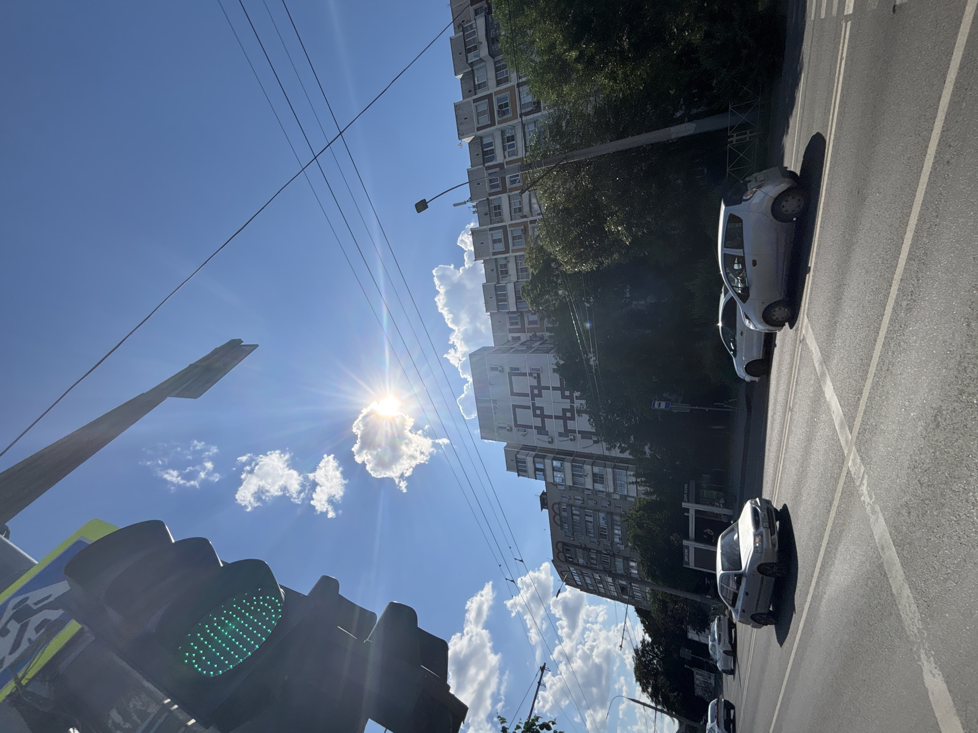 The image depicts a street scene in Krasnodar, Russia, on a sunny day with a clear blue sky and scattered white clouds. A traffic light displaying a green signal is prominent in the upper left corner. In the background, several multi-story apartment buildings line the street, with dense green trees in front of them. Power lines are visible crisscrossing the sky. The street is paved, with lane markings and a pedestrian crossing. Several cars are visible on the road, including a silver sedan in the foreground and a smaller silver hatchback in the middle ground. The sun is shining brightly, casting lens flares and creating shadows on the street. There are no people clearly visible in the image.