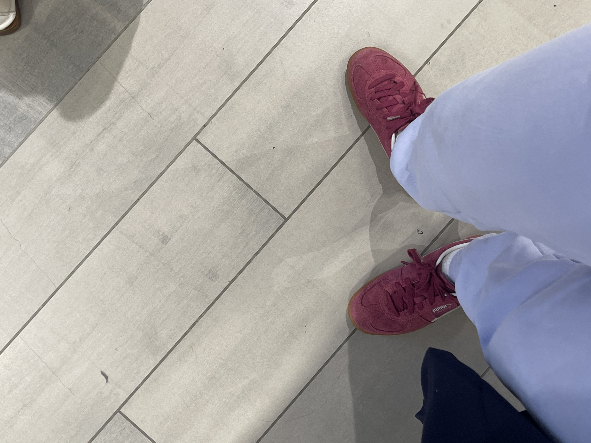 The image captures a high-angle view of a person's legs and feet, standing on a light-colored, possibly off-white or light grey, tiled floor. The floor features rectangular tiles arranged in a linear pattern with grey grout lines, indicating an indoor setting.
The main subject is wearing light blue trousers or sweatpants and a pair of reddish-pink (maroon or burgundy) sneakers. The sneakers have white laces and light brown gum soles. Visible white text on the side of one sneaker clearly reads "PUMA" and "PALERMO", identifying the brand and model. White socks are also visible between the sneakers and the trousers. The person appears to be standing or casually walking, with their feet positioned slightly apart.
In the bottom right corner of the main image, a section of another person's leg, dressed in dark blue trousers, is partially visible. Additionally, a small portion of a light-colored shoe or sandal is visible in the top-left corner. The lighting in the scene is even, suggesting artificial indoor illumination. The setting appears to be a casual public or commercial space, consistent with locations found in Maidstone, United Kingdom, as provided by the context.