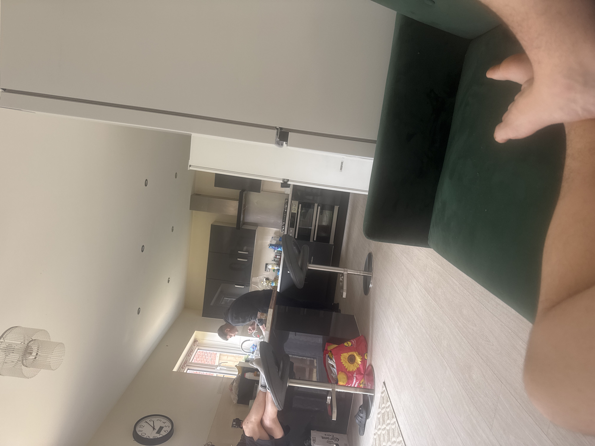The image shows a residential kitchen in Swindon, United Kingdom. A man in a black hooded sweatshirt stands at a black kitchen island, possibly washing dishes or preparing food. Two silver bar stools are positioned at the island. A shopping bag with a sunflower design is visible near one of the stools. In the foreground, a portion of a green velvet couch is visible, along with a partially visible human arm and leg. The kitchen has black cabinets and stainless steel appliances. A clock hangs on the wall, and a window is visible in the background.