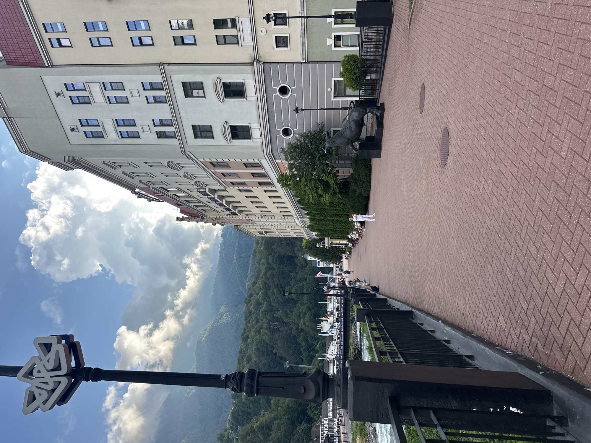 This is a full-shot, eye-level, outdoor, daytime photograph of a waterfront promenade in Estosadok, Russia. The scene features a paved walkway made of red bricks, bordered by a black metal fence on the left and a multi-story building on the right. A river flows along the left side of the walkway, with lush green mountains in the background.
On the left side of the walkway, there is a black lamppost with a decorative white symbol at the top. The building on the right is a modern structure with multiple floors, featuring a facade with various colors and window arrangements. In front of the building, near the walkway, stands a dark-colored statue of a bull.
Several people are present in the scene. A person in white is standing near the bull statue, possibly taking a photo. Other individuals are seated on benches further down the walkway, and some are walking. A bridge crosses the river in the mid-ground. The sky is partly cloudy with patches of blue. The overall atmosphere appears to be peaceful and scenic.