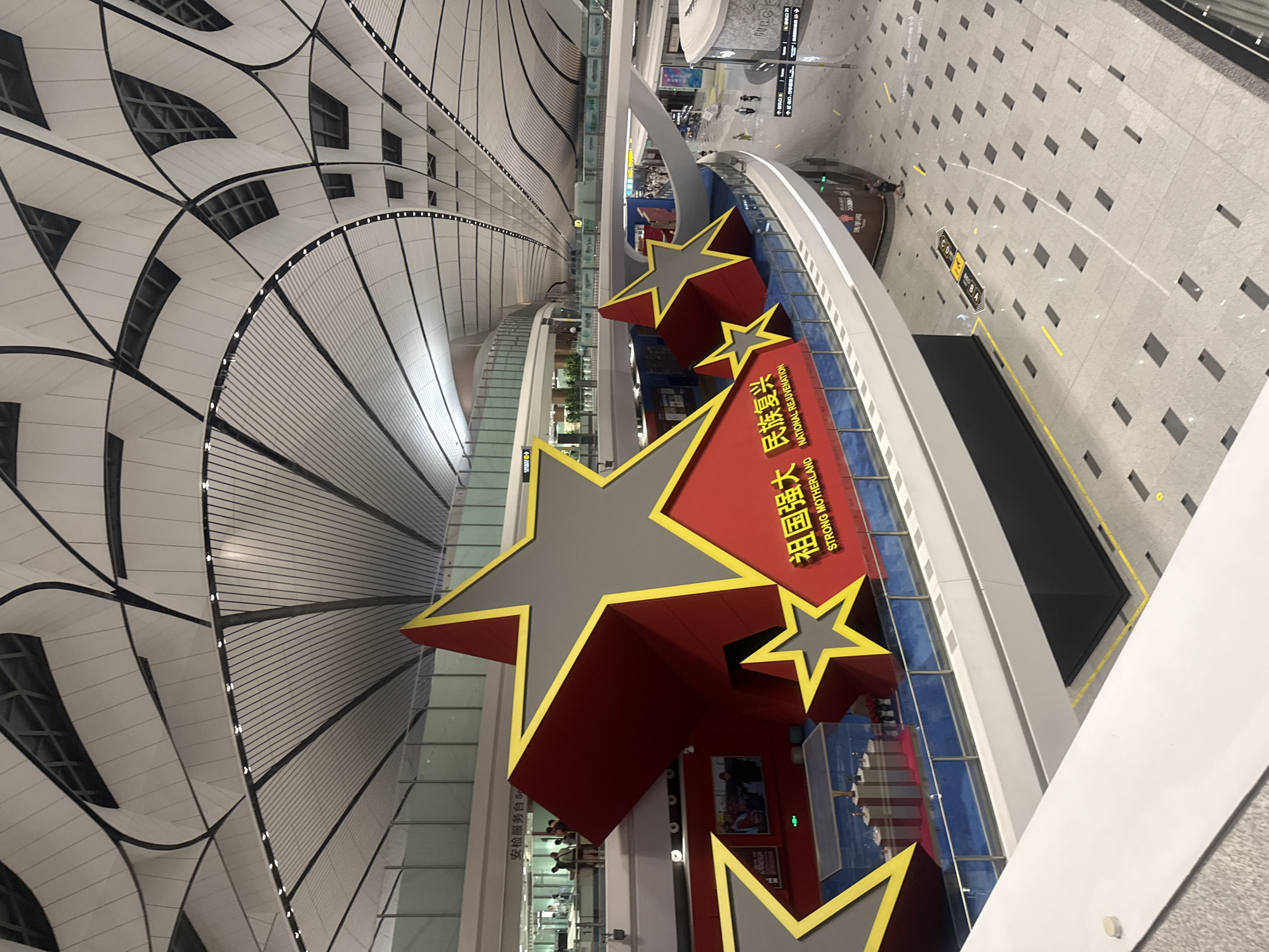 The image shows an interior view of a modern airport terminal. The ceiling is a complex, undulating structure made of white panels and black beams, with rows of small lights. In the foreground, a large red and gray star-shaped sculpture is prominently displayed. The sculpture has yellow trim and is adorned with Chinese characters and the English text "STRONG MOTHERLAND NATIONAL REJUVENATION". Below the main sculpture, there is a display case with a model of buildings. Several people are visible in the background, some appearing to be passengers and others possibly staff. The floor is tiled with a light gray material and features dark square inlays arranged in a pattern. Yellow lines and directional arrows are marked on the floor, indicating pathways. The overall environment is clean, spacious, and contemporary. The location is identified as Guangyang District, China.