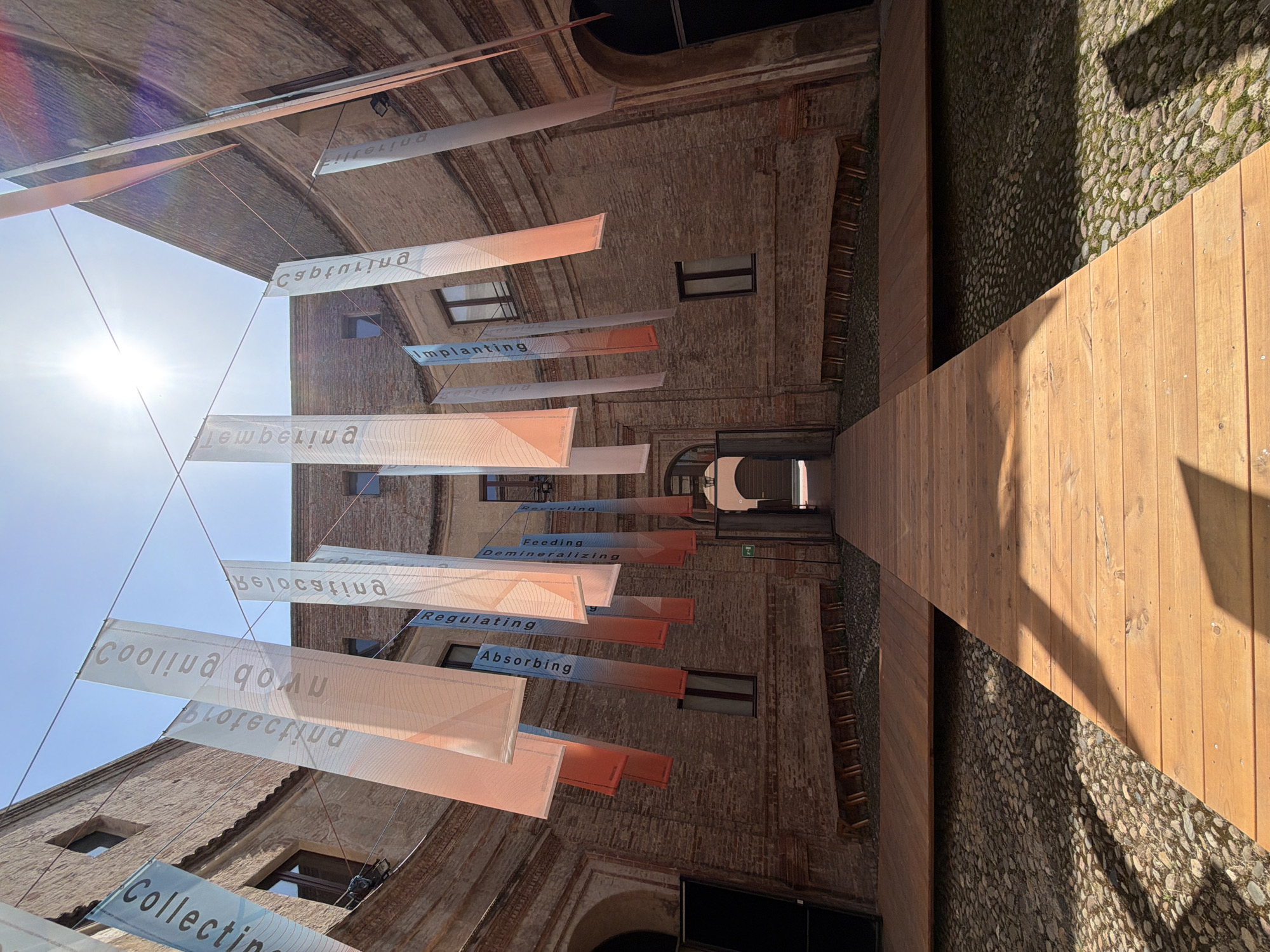 Here is a description of the image:
This is an upward-looking shot in Mantua, Italy, featuring an architectural courtyard with brick buildings on either side. Several translucent banners, suspended by wires, stretch across the courtyard. These banners have words printed on them, including "Filtering", "Transmuting", "Implanting", "Regenerating", "Recycling", "Feeding", "Demineralizing", "Regulating", "Absorbing", "Cooling Down", "Protecting", and "Collecting".
The courtyard floor consists of a wooden pathway that leads to an opening, with pebble-covered ground on both sides of the pathway. The sunlight is visible in the upper left of the frame, adding to the light levels within the courtyard. The buildings display a number of windows of differing sizes, which may indicate architectural details from multiple eras.