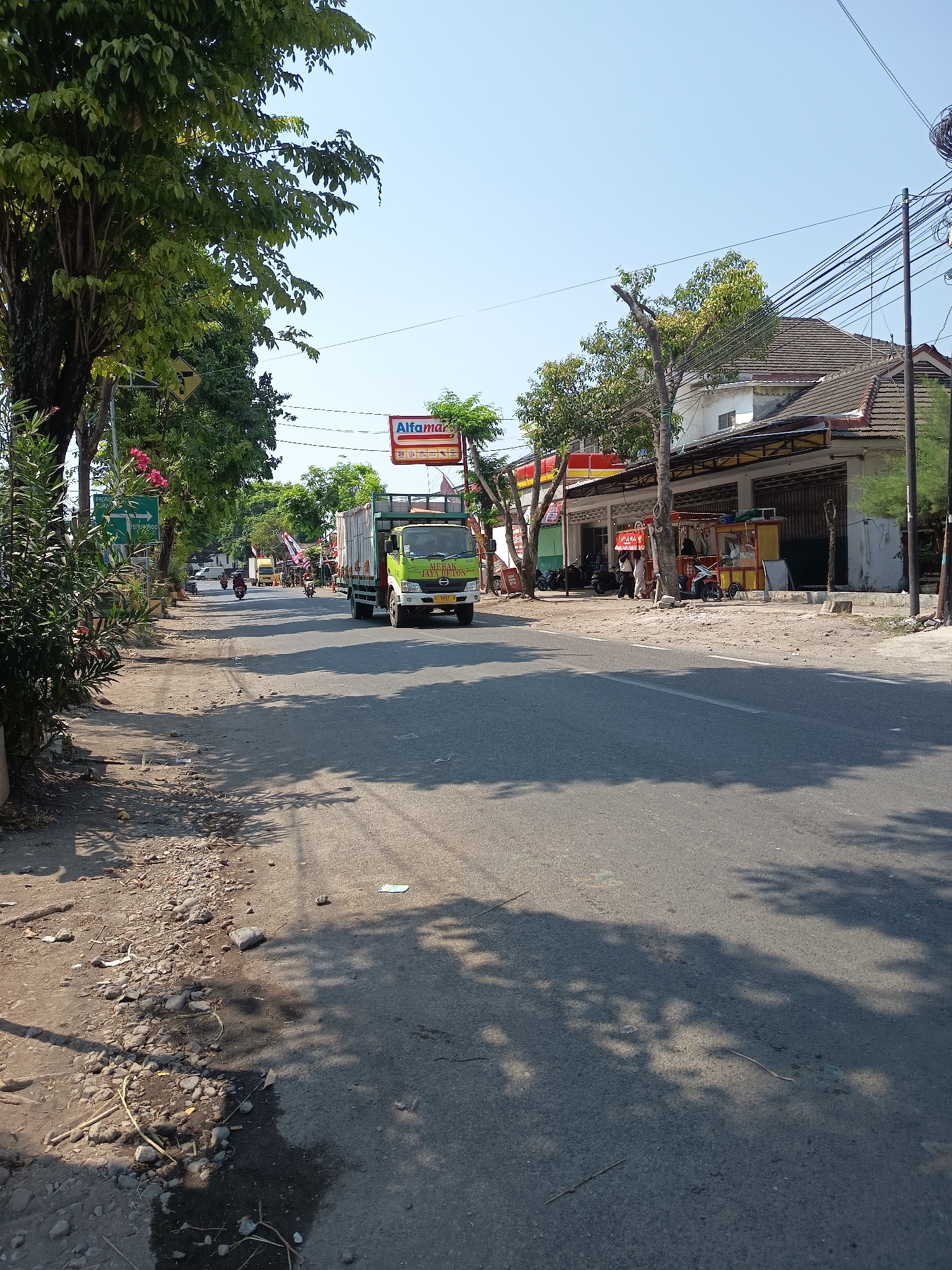 The image depicts a street scene in Kandangjati Kulon, Indonesia, under a clear blue sky. The road is paved and shows signs of wear, with a gravelly shoulder on the left side. Several vehicles are present, including a green and white truck with the text "MORNA JAYATON" on its side, a smaller yellow truck, and multiple motorcycles. A few people are visible, some standing near shops and others on motorcycles.
Along the right side of the road, there are buildings that appear to be shops or small businesses, including an "Alfamart" convenience store with a red and white sign. A food stall with a yellow and red cart is also present. Trees with green foliage line the street, casting shadows on the road. Traffic signs are visible on the left, indicating directions. The overall impression is of a typical, moderately busy street in an Indonesian town.