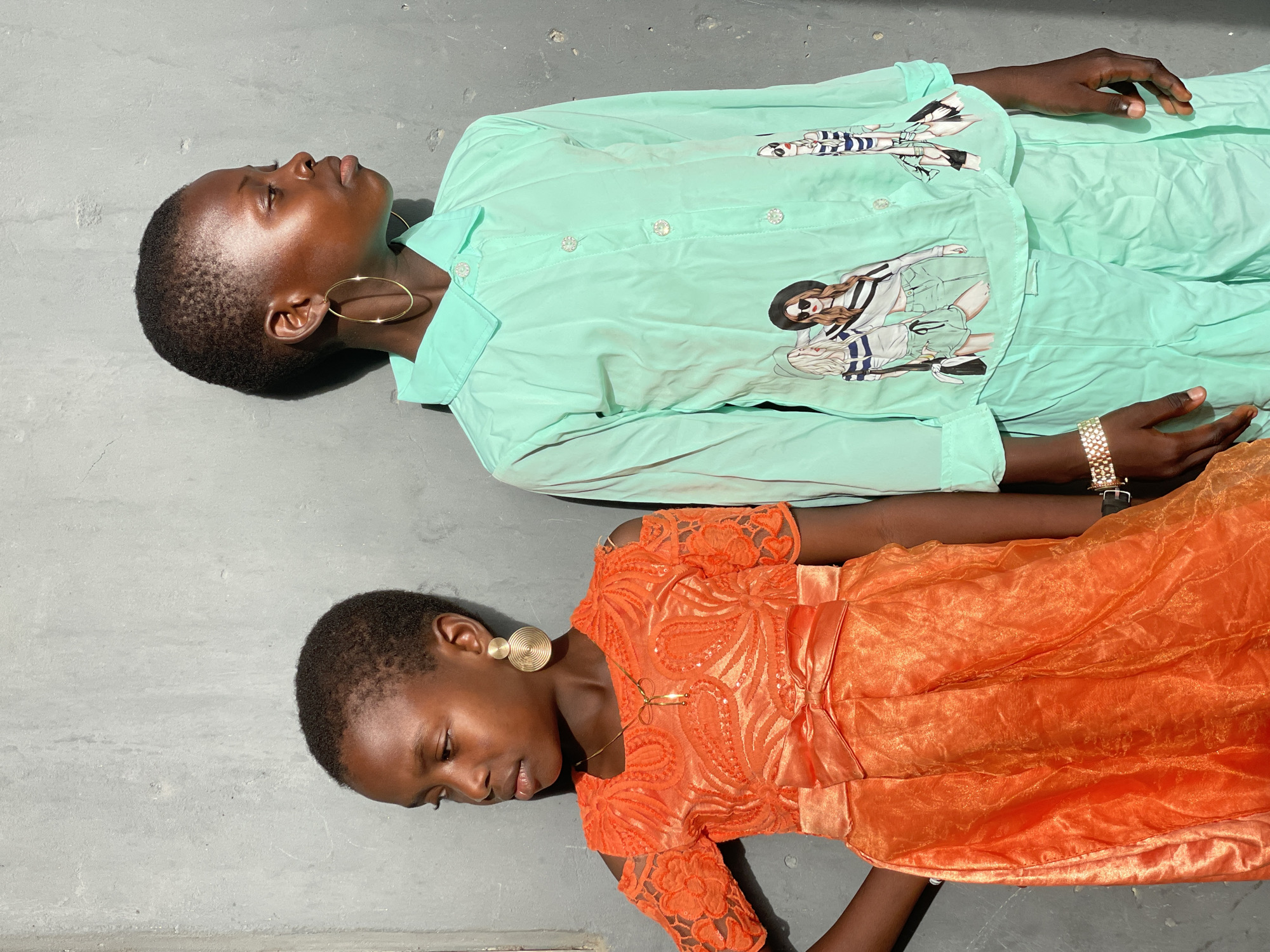 Two young Black individuals are lying on a gray surface. The person on top wears a light green long-sleeved outfit with illustrations. Gold hoop earrings and a gold watch are visible. The person below wears an orange dress and gold earrings. Both appear to be lying on a flat, possibly concrete or tiled surface. The location is implied to be Badagry, Nigeria, but this cannot be confirmed by visual information.