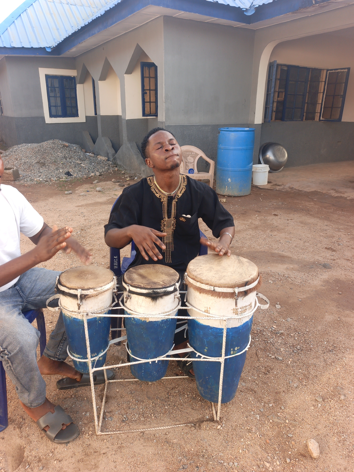 Outside a grey building in Akwanga, Nigeria, two men sit on blue plastic chairs playing conga drums. The drums are white and blue, arranged in a metal stand. A blue barrel, white bucket, and metal bowl sit near the building. The ground is covered in reddish-brown gravel and sand. The man on the left wears a white shirt and jeans; the other wears a black embroidered top.