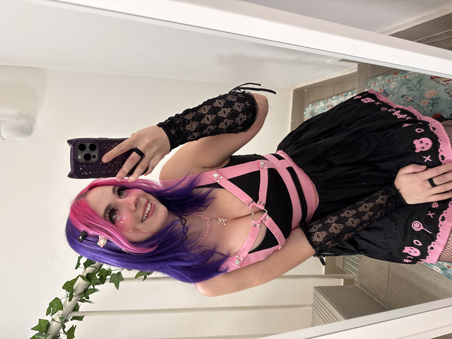 A female subject with two-toned pink and purple hair, adorned with glitter on her face, is depicted taking a mirror selfie. She wears a black sleeveless top with a pink harness-style overlay featuring silver stud and ring details, including a heart-shaped buckle at the sternum. Her attire includes black lace fingerless arm warmers with a diamond pattern and a black skirt with a pink embroidered border showcasing teddy bears, lollipops, 'X's, and 'O's. A black choker with a heart pendant, a pink beaded necklace with a teddy bear charm, and a black ring on her left index finger are visible. She holds a dark-colored smartphone, encased in a cat-ear design, in her right hand (viewer's left). The background consists of a plain white wall, a white railing with artificial green ivy on the left, grey steps, and a partially visible patterned rug on the floor. The environment is an indoor setting. No specific visual cues are present within the image to identify a precise location within the City of Zagreb, Croatia.