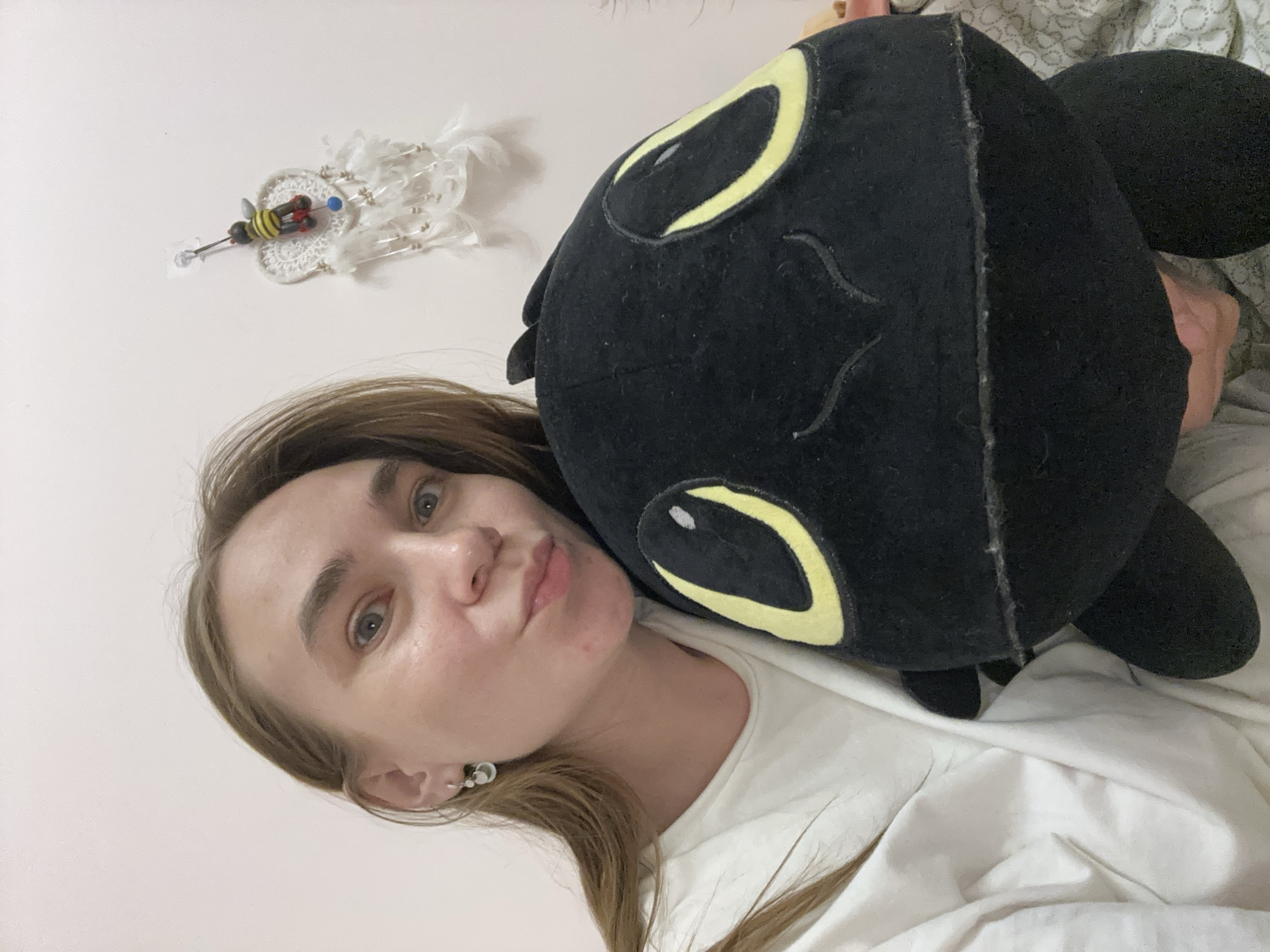A woman with light brown hair and light-colored eyes is taking a selfie with a black dragon plushie. She is wearing a white t-shirt and small, silver earrings. The background includes a white wall with a decorative item hanging on it, and a glimpse of patterned bedding. The location is not identifiable as the city of Kharnavy, Russia from this image alone.