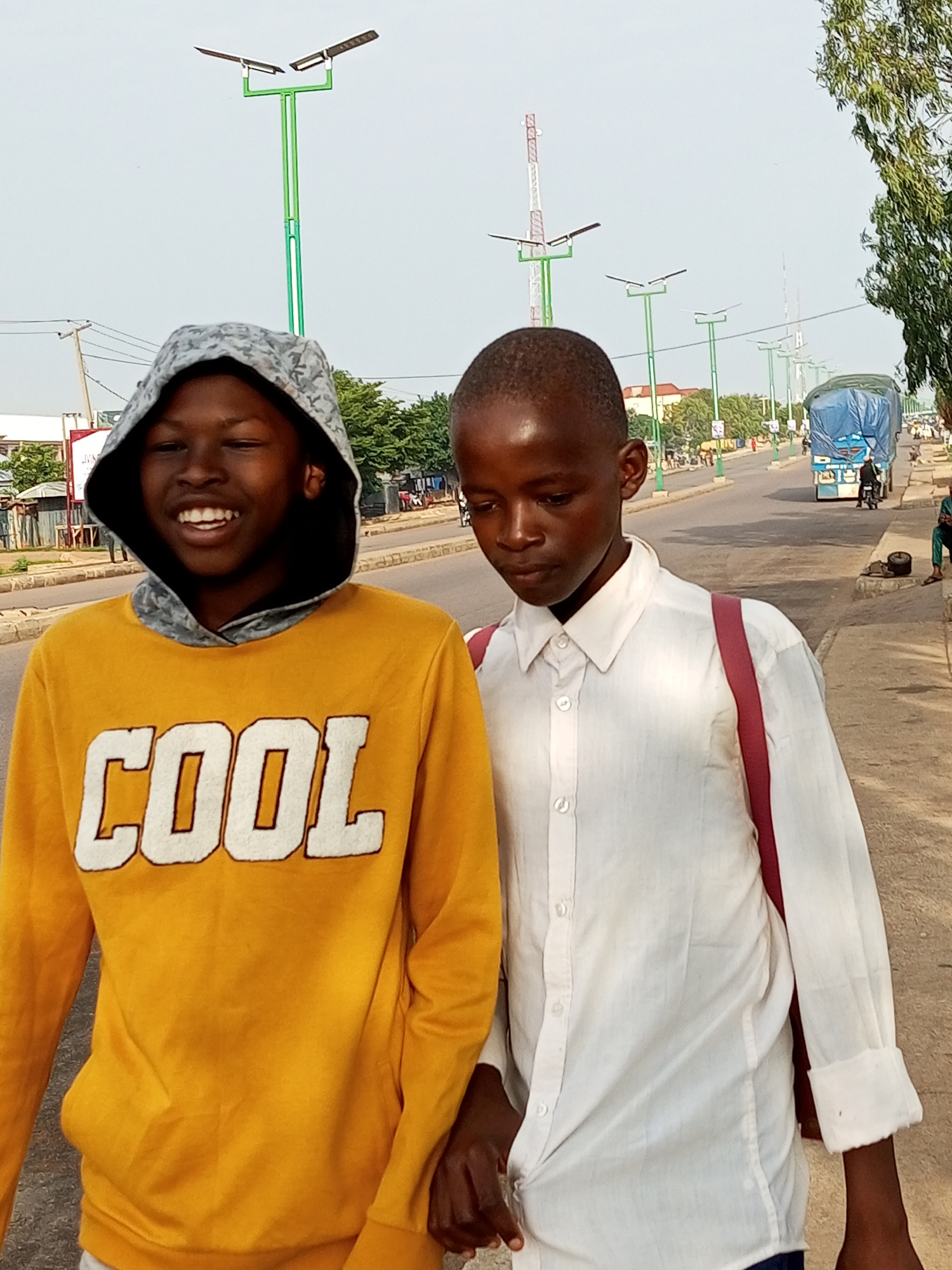 Two young boys are standing on a paved road in Tumfure, Nigeria. The boy on the left is wearing a yellow hooded sweatshirt with the word "COOL" printed in white letters. He is smiling and looking at the camera. The boy on the right is wearing a white button-down shirt and a maroon backpack. He is looking down. In the background, there are streetlights, a blue truck, and other vehicles. The sky is overcast.