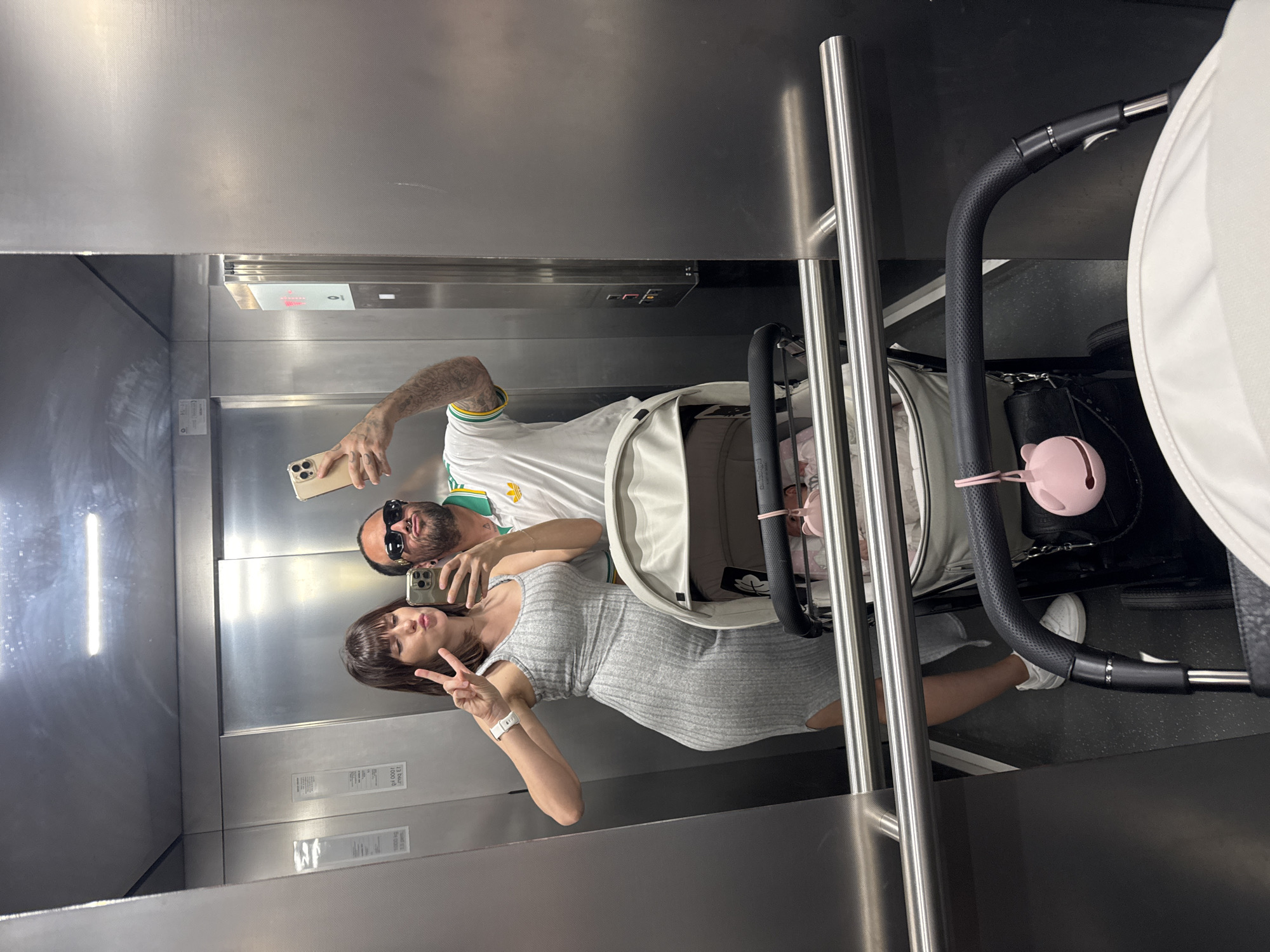 In an elevator interior, a man and a woman are visible in a mirror taking a selfie. The man, with a beard and wearing sunglasses, is holding a smartphone in his left hand. He is wearing a white t-shirt with green and yellow accents. The woman, wearing a grey dress, holds up two fingers in a peace sign. A stroller occupies the right side of the elevator. The elevator's metal interior is visible with control panels and signage on the walls. The location is in Pomezia, Italy, based on signage visible in the mirror.