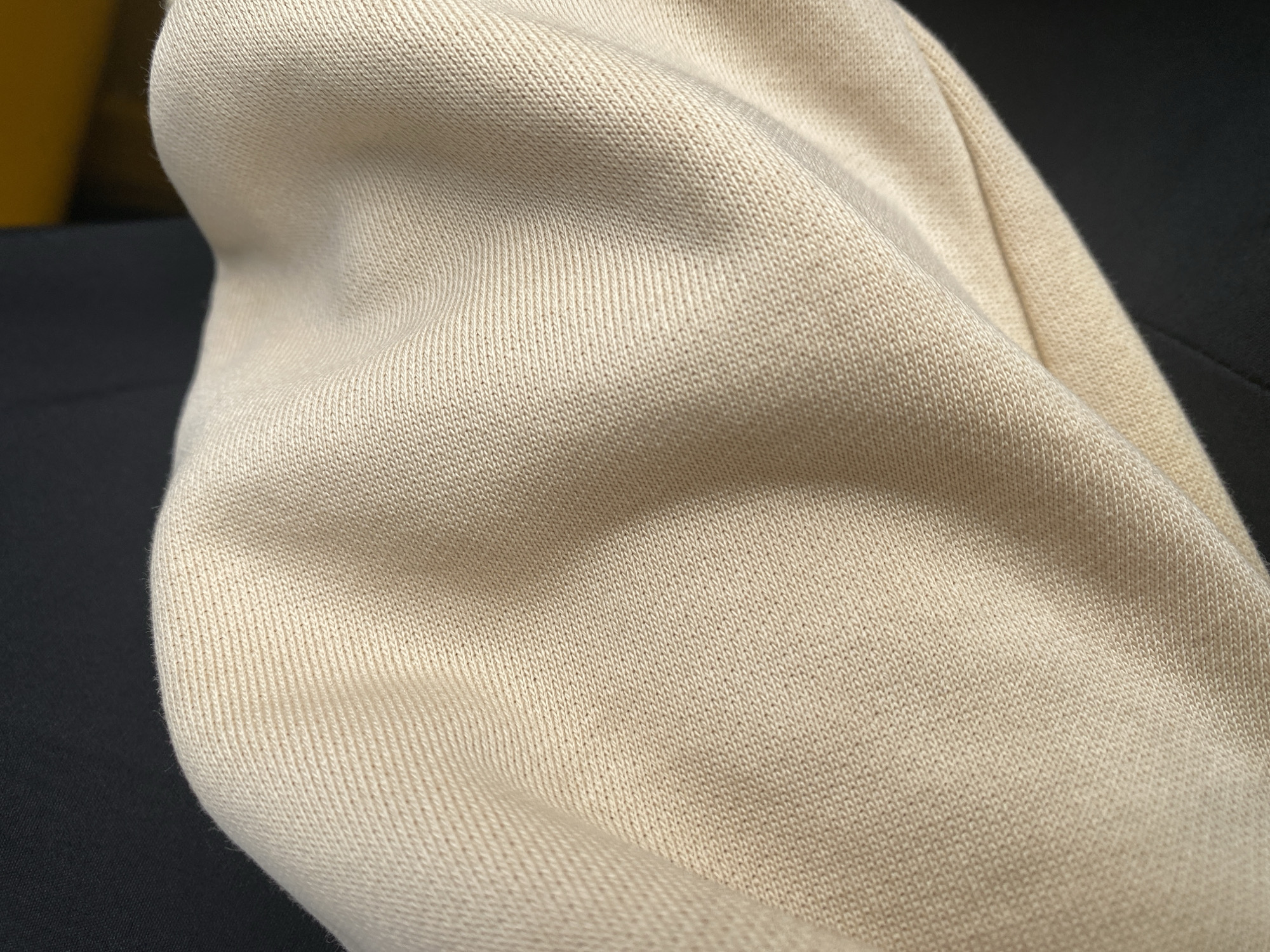 The image is a close-up, top-down view of a light beige knitted fabric, likely a sweater or sweatshirt, draped over a dark surface. The fabric exhibits subtle wrinkles and folds, creating soft shadows that highlight its texture. The knit pattern is visible, characterized by small, interlocking loops. In the upper left and lower right of the frame, a dark, out-of-focus background is present, possibly clothing or furniture. The lighting appears to be natural, casting gentle shadows across the fabric's surface. There are no people or identifiable landmarks visible, making it impossible to determine a specific location within Jalingo, Nigeria or anywhere else. The scene is static and focuses solely on the texture and form of the textile.