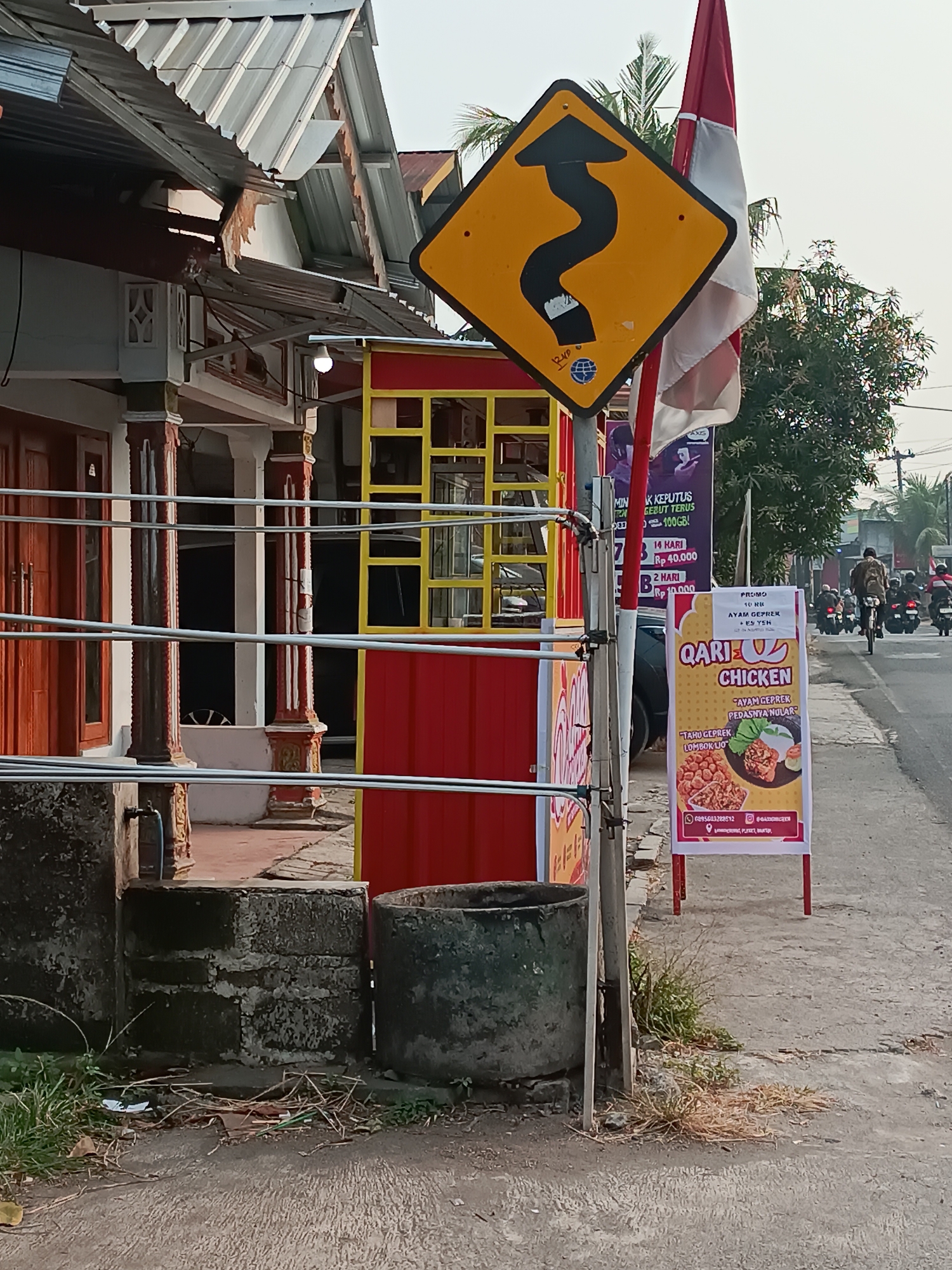 The image depicts a street scene in Pleret, Indonesia, with a focus on a roadside stall and traffic signs. In the foreground, a winding road sign warns of curves ahead, with a red and white Indonesian flag partially obscuring it. To the left, a building with red doors and decorative pillars suggests a residential or commercial establishment. A red and yellow food stall is visible behind the sign. A "Qari Chicken" advertisement poster displays images of fried chicken and rice. In the background, several motorcycles are seen on the road, indicating ongoing traffic. The overall environment appears to be an urban or semi-urban setting, with some vegetation and weathered structures.