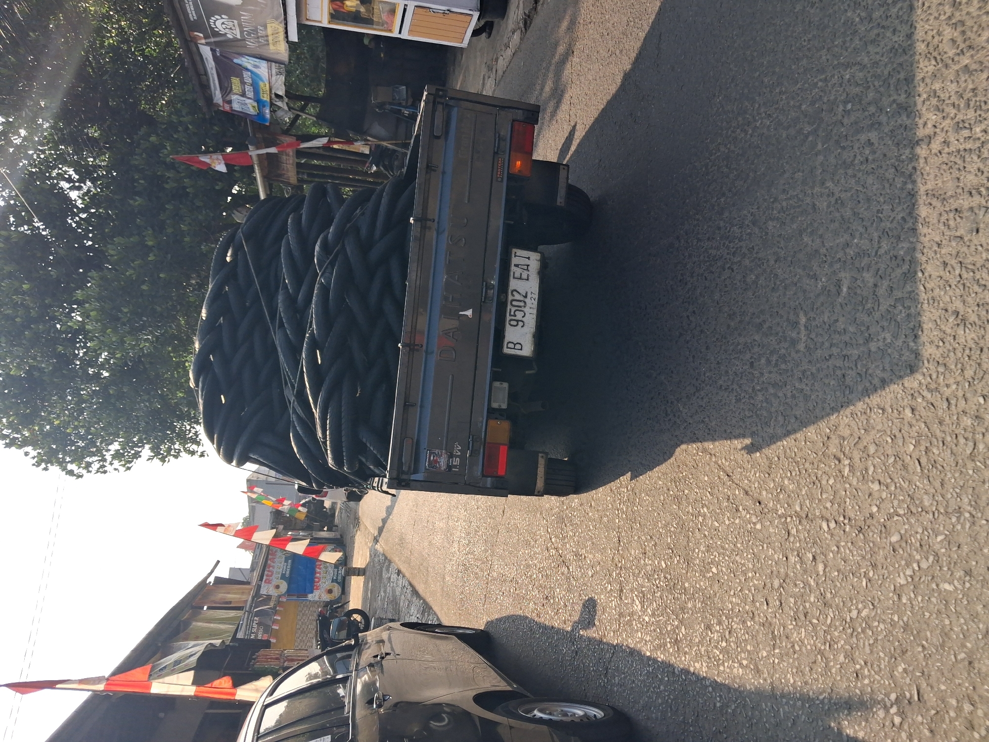 In Bojong Gede, Indonesia, on a sunlit street with asphalt paving, a dark gray pickup truck is prominently featured, loaded with a large quantity of black tires. The truck displays a license plate reading "B 9502 EAI" and the "DAIHATSU" logo on its tailgate. To the left of the truck, the rear passenger side of a black vehicle is visible, casting a shadow on the road. The surrounding environment includes buildings with red and white Indonesian flags displayed, suggesting a possible national celebration. Trees line the background, and a food stall with a yellow and white structure is present on the right side of the frame. The scene depicts a street, possibly in a commercial or residential area, during daylight hours.