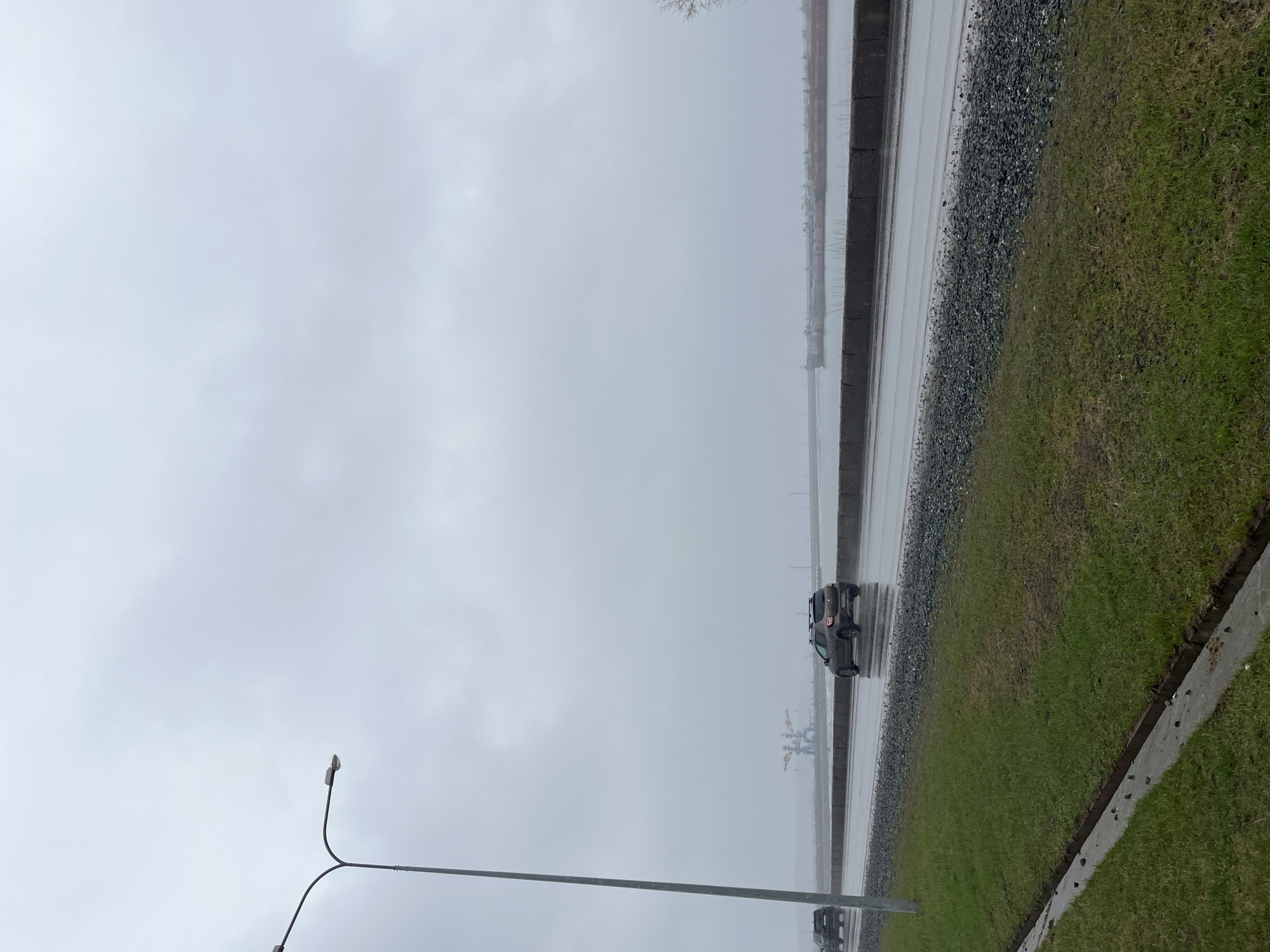 An outdoor, landscape-oriented photograph captures a gloomy, overcast day in what appears to be an industrial or waterside area. The scene is dominated by a wide, uniformly light grey sky, suggesting heavy cloud cover, possibly with rain or mist.
In the foreground on the left, a street light pole with two upward-curving lamps is prominently featured. Below it, a strip of vivid green grass borders a concrete curb, which separates it from a dark, wet paved surface—likely a road. Adjacent to the road is a narrow band of dark gravel or small stones, followed by another concrete barrier or edge.
A dark-colored SUV or car is seen driving on the wet road, moving away from the viewer. Its surface appears damp, reflecting the light from the sky. Another vehicle is partially visible in the distance, behind the street light pole.
In the hazy background, indistinct industrial structures, possibly cranes or other port machinery, are faintly visible, suggesting the presence of a harbor, port, or large body of water. The entire scene is wet, with the road surface reflecting the overcast sky, indicating recent or ongoing rain.
The time of day is daytime, but the heavy clouds and wet conditions lend a somber and muted atmosphere to the environment. No discernible text is visible in the image.