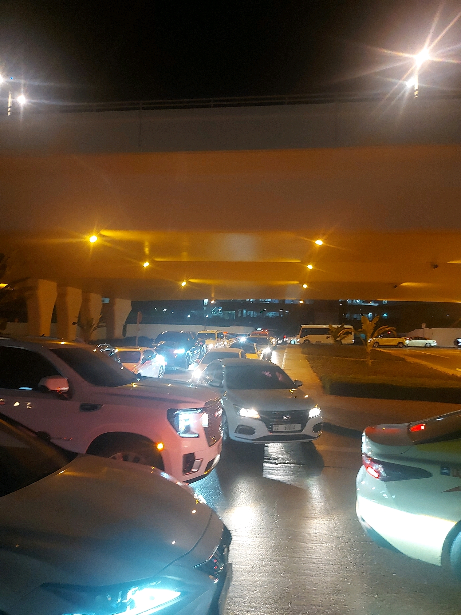 The image shows a nighttime traffic jam on a multi-lane road in Dubai, United Arab Emirates. The road is wet, reflecting the bright overhead lights and vehicle headlights. Numerous cars are stationary or moving slowly in dense traffic, indicating congestion. On the right, a white sedan with its taillights illuminated is visible. In the center, a white Hyundai sedan is also present, its headlights on and a license plate reading "33 51014" is visible. A pinkish-white SUV is in the foreground on the left. Above the road, a large overhead structure, likely a flyover or bridge, casts a warm orange glow from its lighting. The dark sky and illuminated signs suggest an urban environment. No people are clearly visible in the frame. The scene captures a common urban traffic scenario at night in Dubai, characterized by heavy vehicle presence and artificial lighting.