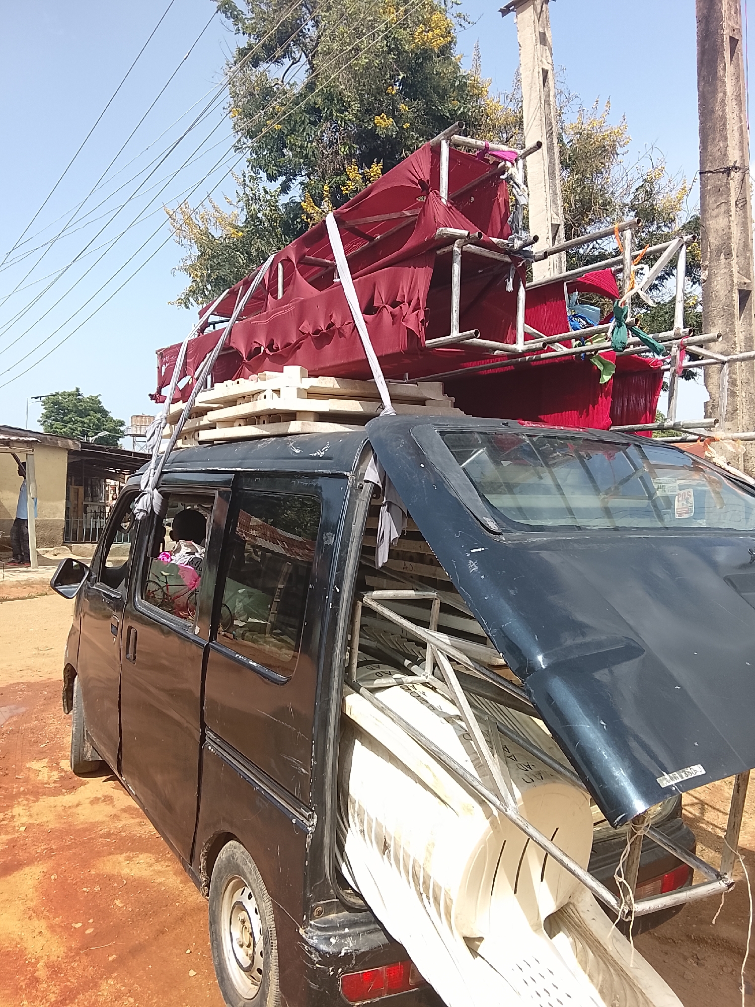 A black van, heavily laden with scaffolding, red fabric, and what appear to be white wooden structures, is parked on a dirt road in front of a building. The scaffolding is stacked high on the roof of the van, with red fabric draped over parts of it. Several white, rectangular objects, possibly furniture components or building materials, are also visible on the van and precariously balanced on the scaffolding. The van's rear door is open, revealing more of the white structures inside. A person, partially obscured by the van's window, is seated in the driver's seat. The environment appears to be an outdoor, possibly rural or semi-urban setting, with trees, overhead wires, and utility poles visible in the background. The ground is covered in reddish-brown dirt. The sky is clear and blue.