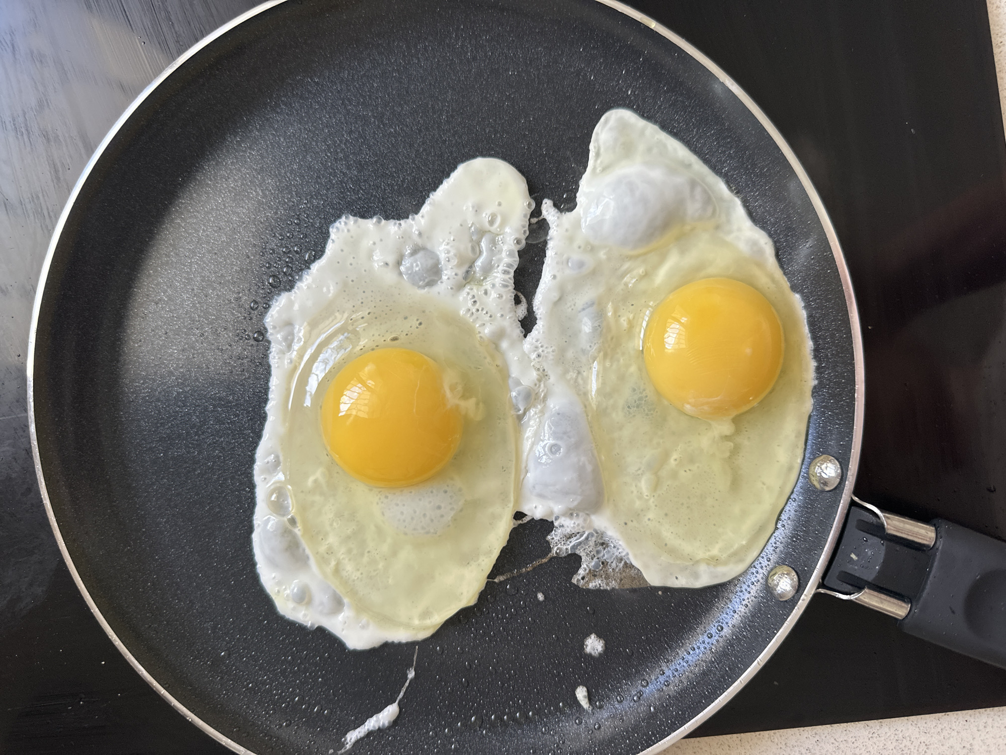 The image shows two sunny-side-up eggs frying in a black non-stick pan. The yolks are bright yellow and the whites are mostly cooked, with some bubbly and translucent areas. The pan is on a flat black stovetop surface. A black pan handle extends out of the bottom right corner. The image was taken indoors, under adequate lighting. There is no evidence in the image to suggest a location in the city of Petah Tikva, Israel, or any other specific location. No people are visible.
