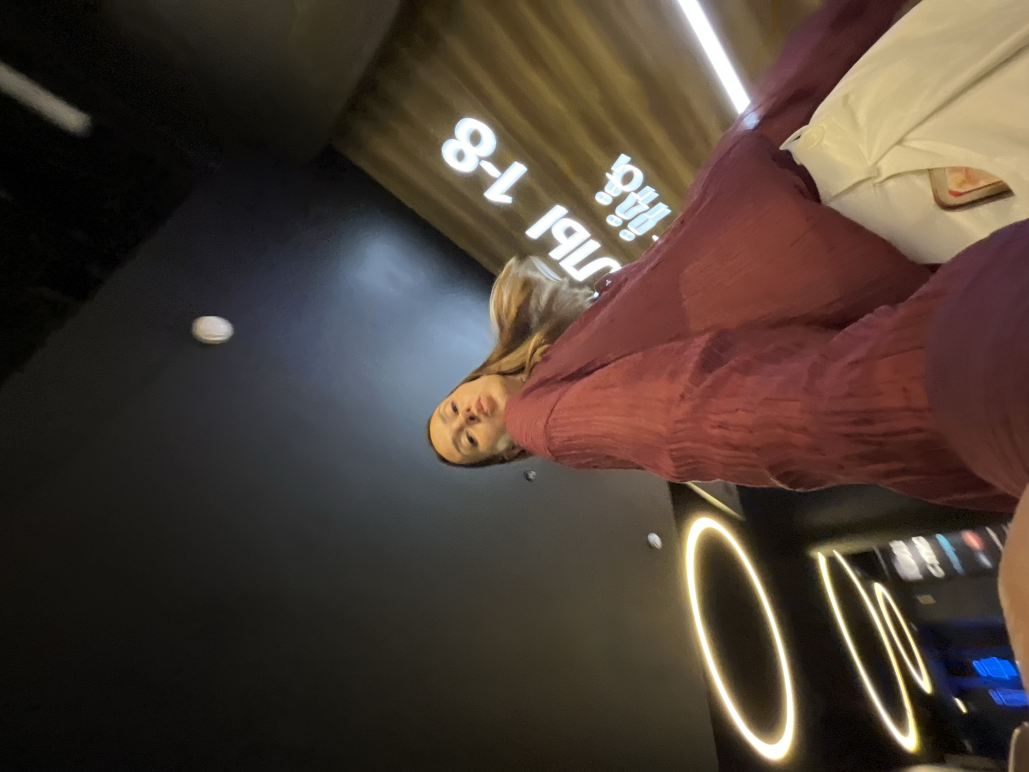 A low-angle, wide shot captures a young woman in a dark red, crinkled long-sleeved shirt and white pants posing for a selfie in what appears to be a dimly lit cinema lobby or hallway in Sochi, Russia. She is positioned in the foreground, looking directly at the camera with her lips pursed. Her blonde hair is slightly blurred, suggesting movement. In the background, wooden-paneled walls display illuminated signs. One sign prominently features white, glowing Cyrillic text that reads "РЯДЫ 1-8" (Rows 1-8). Below this, smaller illuminated icons are visible. Circular, yellow-white LED lights are mounted on the dark ceiling, creating a halo effect. The overall ambiance is modern and theatrical, consistent with a cinema environment in Sochi.