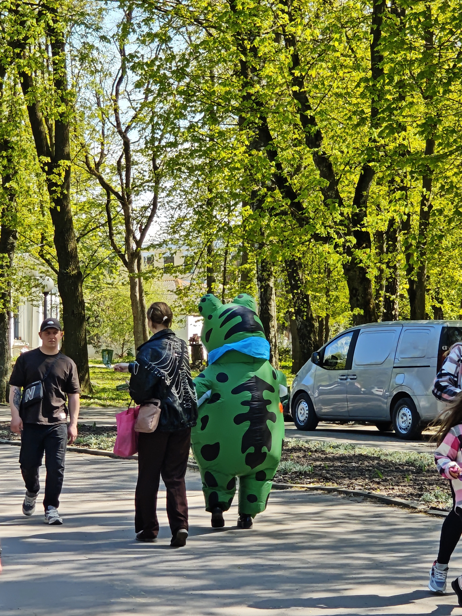The image captures a scene on a sunny day in a park, likely in Vinnytsia, Ukraine. A man wearing a black baseball cap and t-shirt walks from left to right. Next to him, a woman with a black jacket and brown trousers stands next to a person wearing a green dinosaur costume with black spots and a blue bandana. A grey van is parked nearby. Trees with green leaves form a backdrop for the scene. A girl wearing a pink flannel can also be seen on the right.