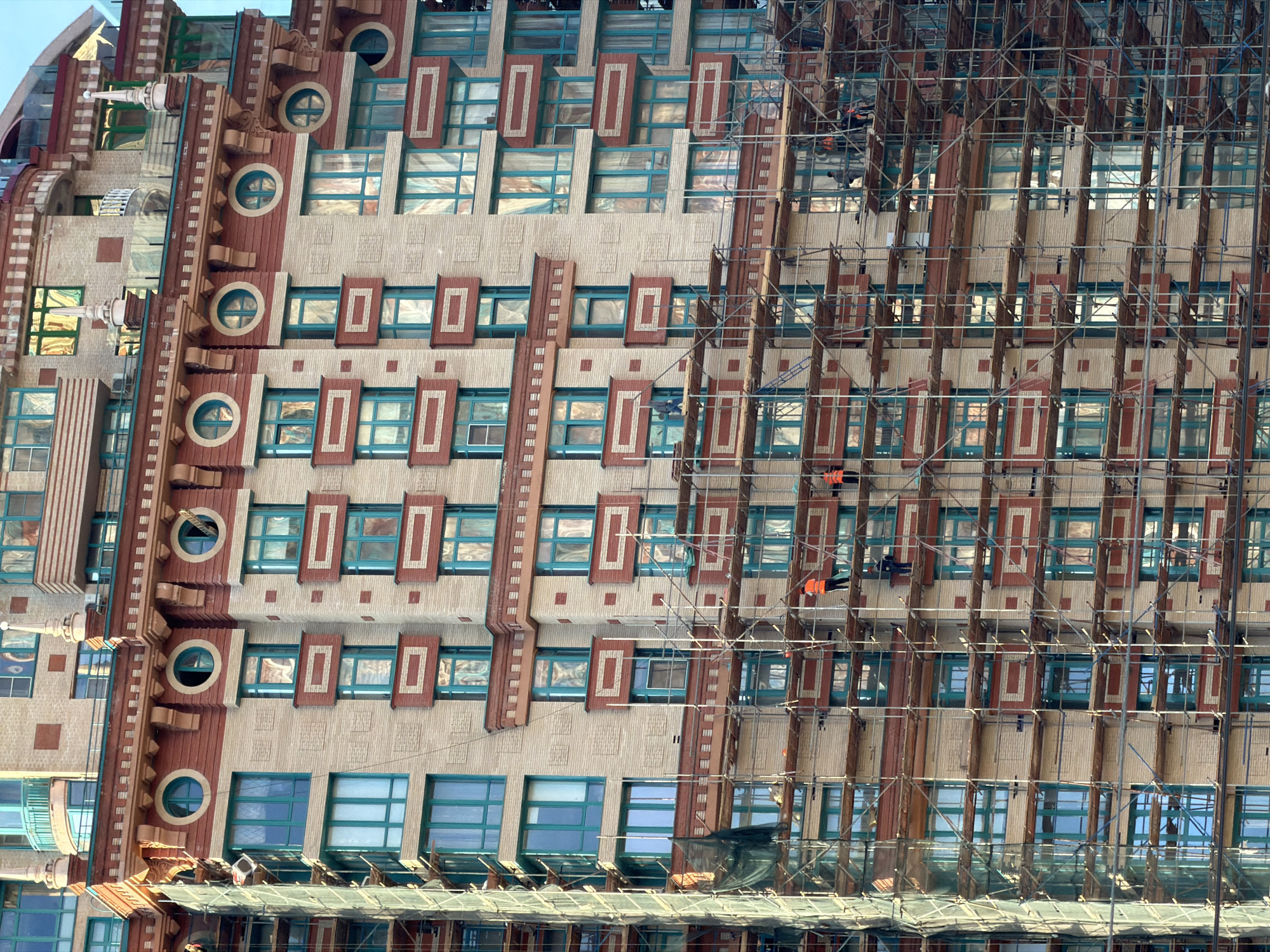 The image depicts the exterior of a building under construction or renovation, with scaffolding partially obscuring the facade. The building features a repetitive pattern of rectangular windows with teal frames, accented by vertically-oriented, reddish-brown decorative elements. On the left side, there's an additional section with circular windows and more intricate detailing. Two workers wearing orange safety vests are visible amidst the scaffolding. The location is described as the city of Khimki, Russia.