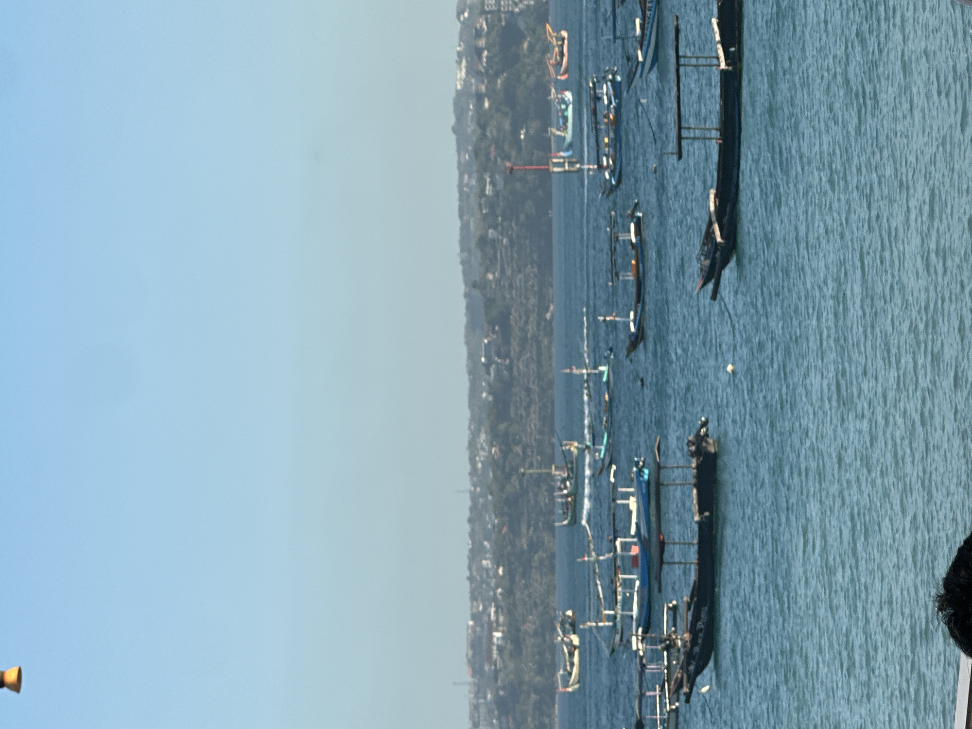 A wide shot shows the Kuta beach in Bali, Indonesia, with numerous fishing boats of various sizes bobbing on the turquoise sea under a clear blue sky. The boats are mainly traditional Jukung-style outrigger canoes, many with canopies for shade. Some boats are anchored, while others appear to be in motion. In the background, a densely populated coastline is visible, lined with buildings and lush greenery, indicating the urban sprawl of Kuta. The water exhibits gentle ripples, suggesting calm weather conditions. The scene captures a typical day at this popular Indonesian beach, a hub for fishing and tourism.