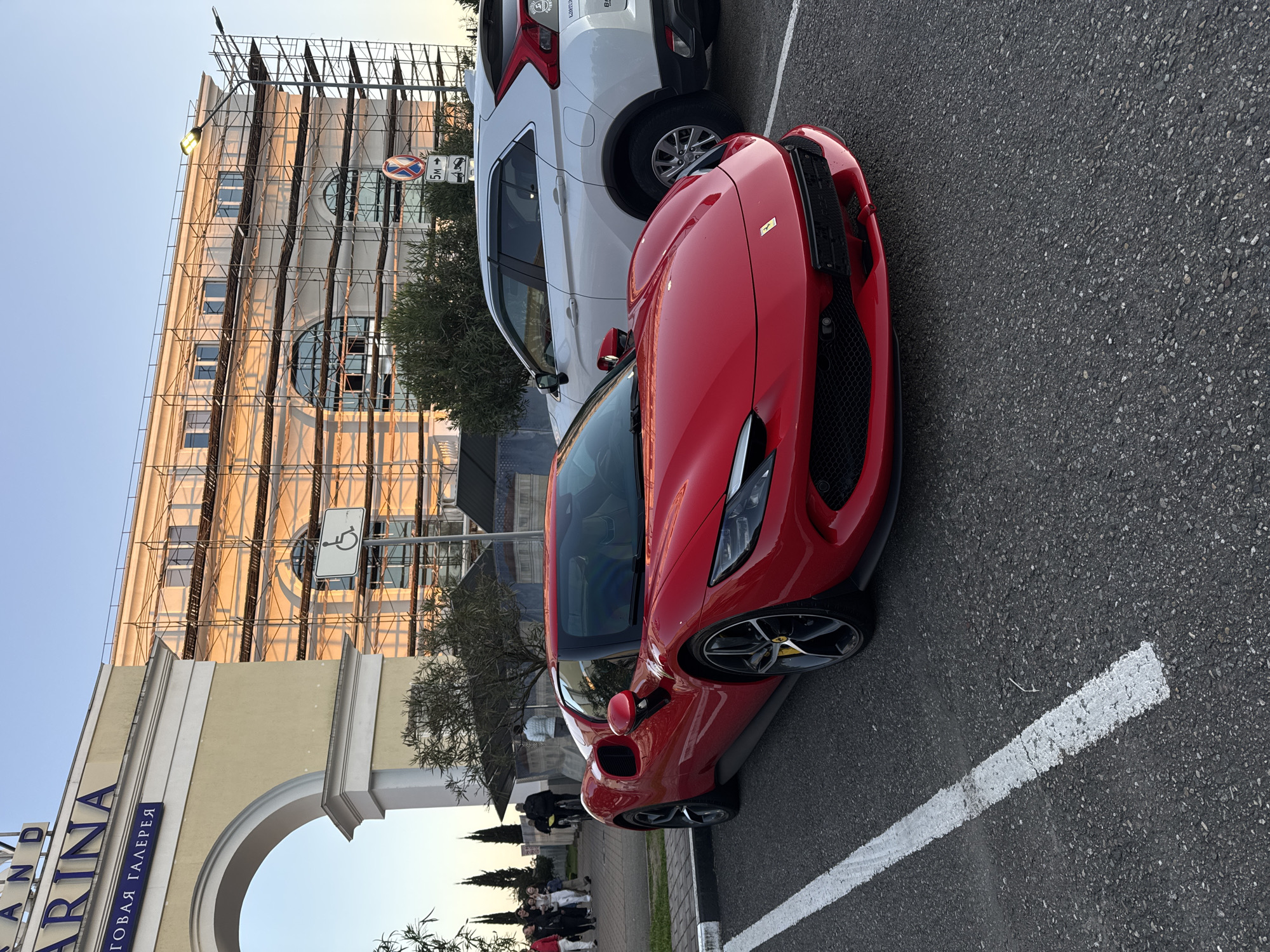 The scene captures a red Ferrari parked in a parking space marked by a white line. Beside the Ferrari is a white SUV. The setting includes a building with scaffolding, a handicap sign, and an arched entrance bearing the inscription "MARINA." A group of individuals is visible in the lower-left corner of the frame. The location is an urban environment.