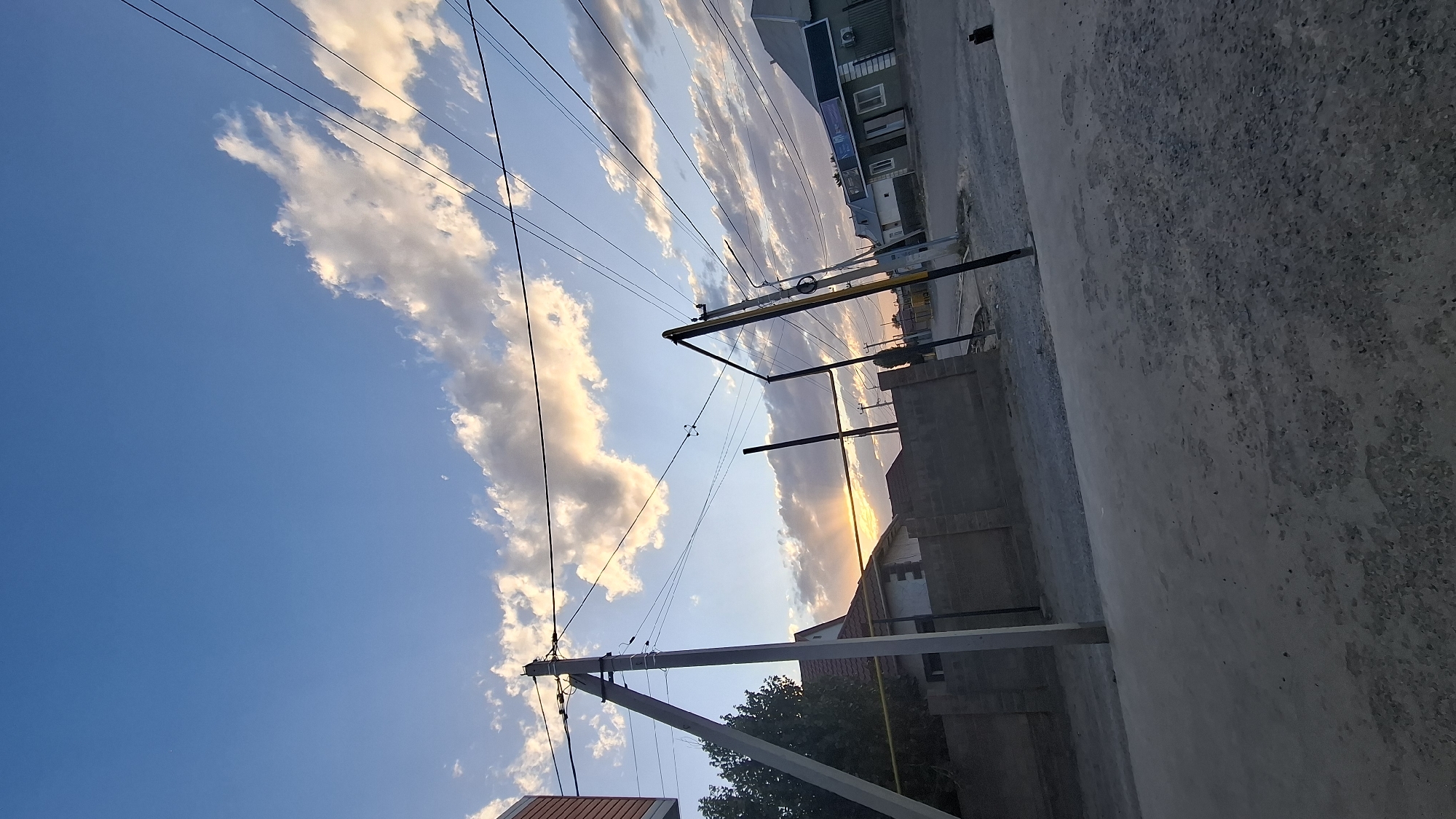 The image depicts a street scene in the settlement of Транспортник, Kazakhstan, under a partly cloudy sky at dusk. Power lines stretch across the frame, supported by concrete utility poles. In the foreground, a gravel road is visible. To the left, a concrete fence encloses a property with a house featuring a red-tiled roof. Further down the street, a commercial building with signage can be seen on the right. The setting sun casts a warm glow, with rays of light visible through the clouds. No people are present in the image.