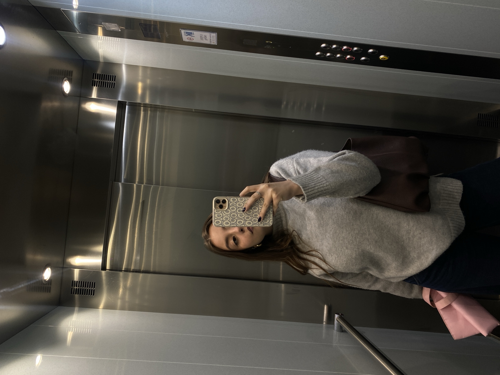 A woman is taking a selfie in an elevator. The elevator has stainless steel walls and doors, with a panel of buttons above the doors. The woman is wearing a gray sweater and dark pants, and she has a brown bag slung over her shoulder. She is holding a phone with a patterned case in her left hand. A pink object is also visible to the right of the scene. The location is Unknown, Unknown.