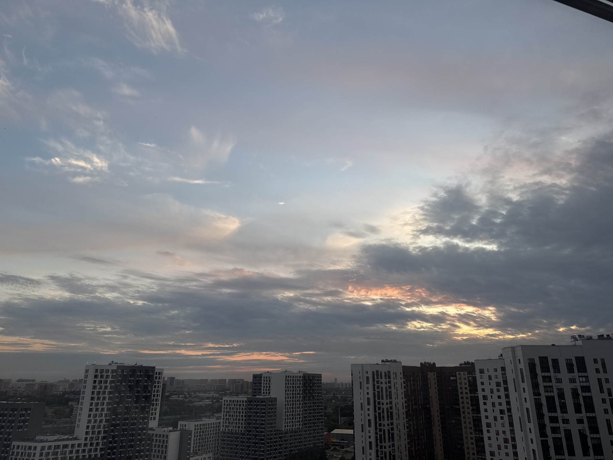 The image depicts a panoramic view of the city of Lyubertsy, Russia, during what appears to be either sunrise or sunset. The sky is filled with dramatic clouds, ranging in color from soft blues and grays to warmer oranges and pinks where the sunlight filters through. Several modern, high-rise apartment buildings with distinct architectural features, characterized by their modular window designs and varying facade colors (primarily white, gray, and dark brown), dominate the foreground and mid-ground of the scene. The buildings are densely packed, suggesting a residential urban environment. In the background, a sprawling cityscape with lower-rise buildings and hints of greenery can be observed stretching towards the horizon, partially obscured by atmospheric haze. No people are visible in the image, and the scene is static, focusing solely on the architectural landscape and the atmospheric conditions of the sky. The overall impression is one of a contemporary urban setting under a dynamic sky.