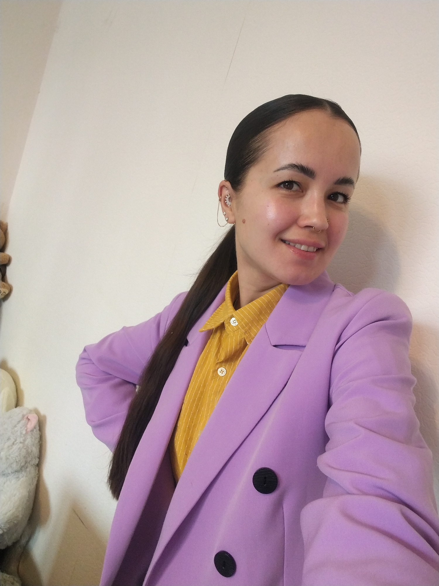 A woman with dark hair pulled back in a ponytail, wearing a yellow shirt and a light purple blazer, stands against a white wall. She is smiling at the camera. She has piercings in her ear and nose. To the left, partially visible, are stuffed toys. The location is not mentioned or indicated in the picture.