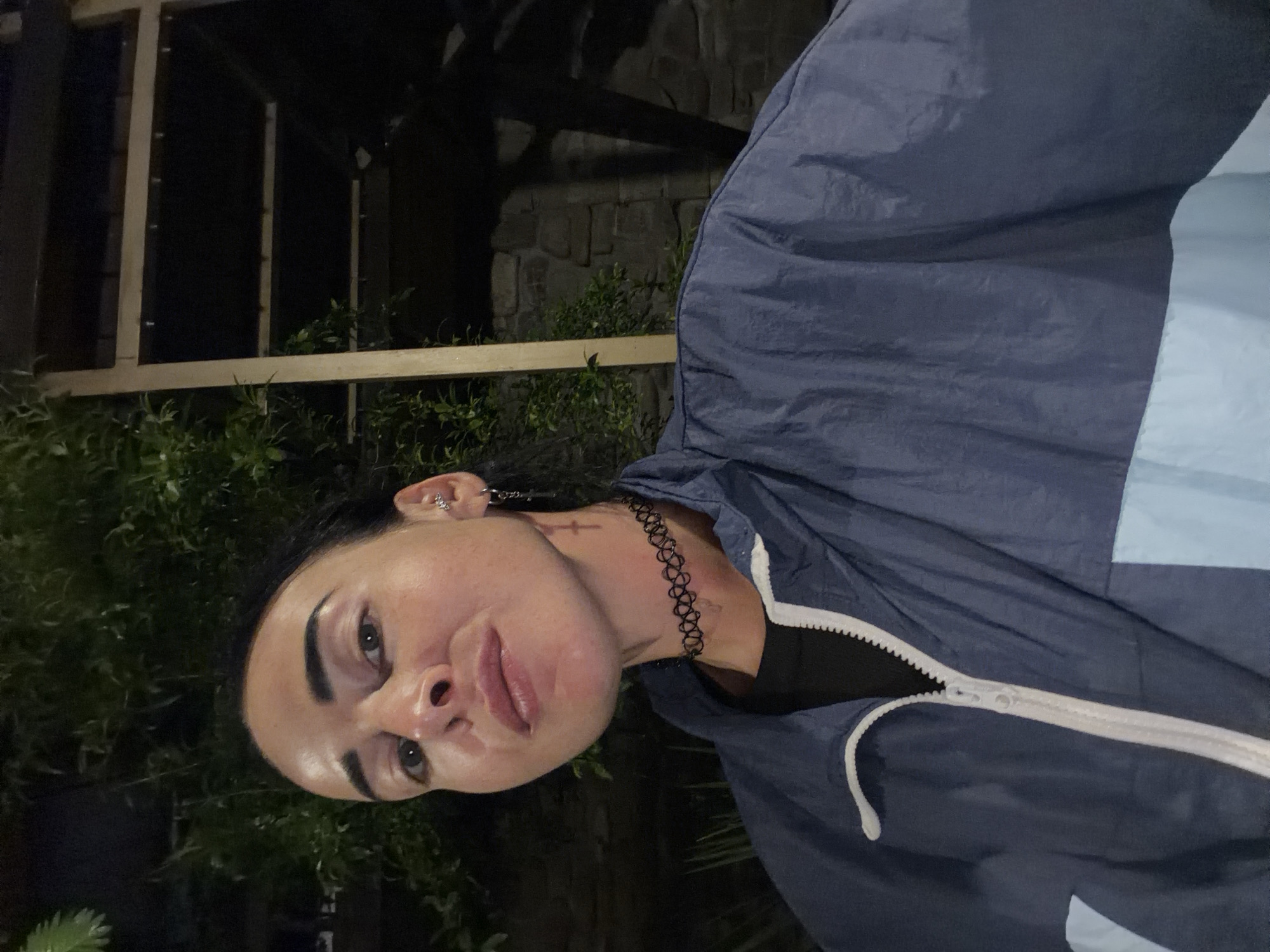 In the provided image, a woman is seen in a close-up shot, with her upper body and head prominently featured. She has dark hair pulled back and is wearing a dark-colored, possibly black, t-shirt underneath a navy blue and light blue zip-up jacket. A black choker necklace is visible around her neck, and she has a cross tattoo on her neck. She also has piercings in her ears and a small earring with a dangling cross. Her facial expression is directed towards the camera, with her lips slightly parted. The background appears to be outdoors at night, with foliage and what looks like a stone wall and wooden structure visible. The lighting suggests artificial light sources. The image quality is slightly grainy due to low light conditions. The specific location within Sochi, Russia, is not discernible from the visual information provided in the image.