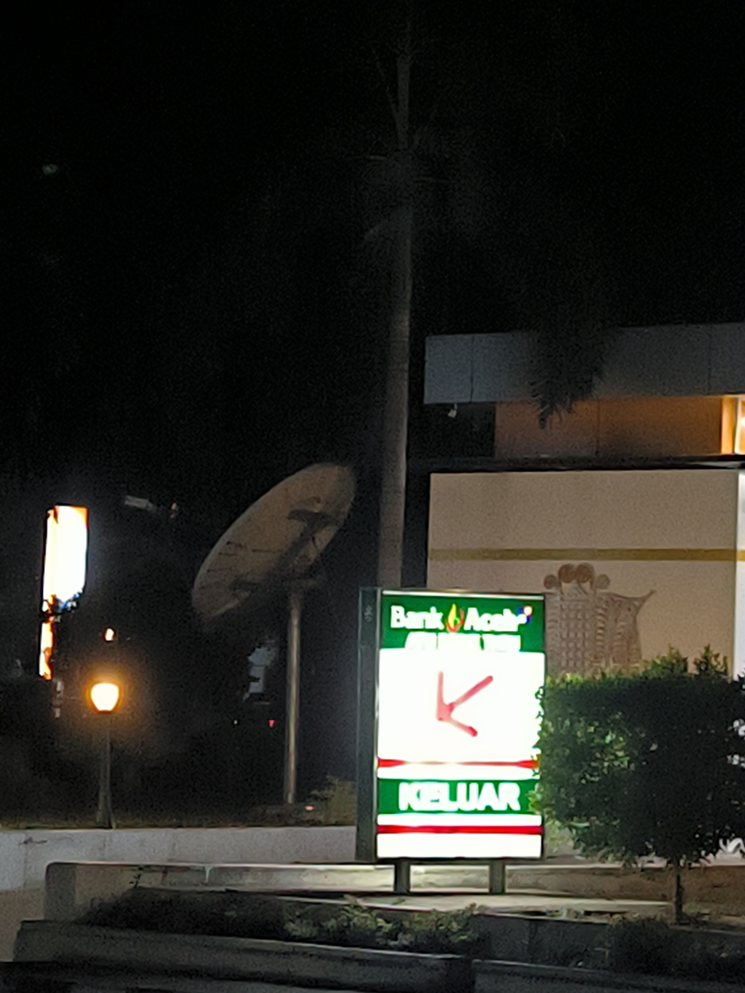 The scene depicts a nighttime view in Lhokseumawe, Indonesia. A sign for "Bank Aceh" is prominently displayed, with a red arrow pointing left above the word "KELUAR". Behind the sign, there is a satellite dish and a building. A street lamp illuminates the foreground. The surrounding environment includes vegetation and a dark sky.