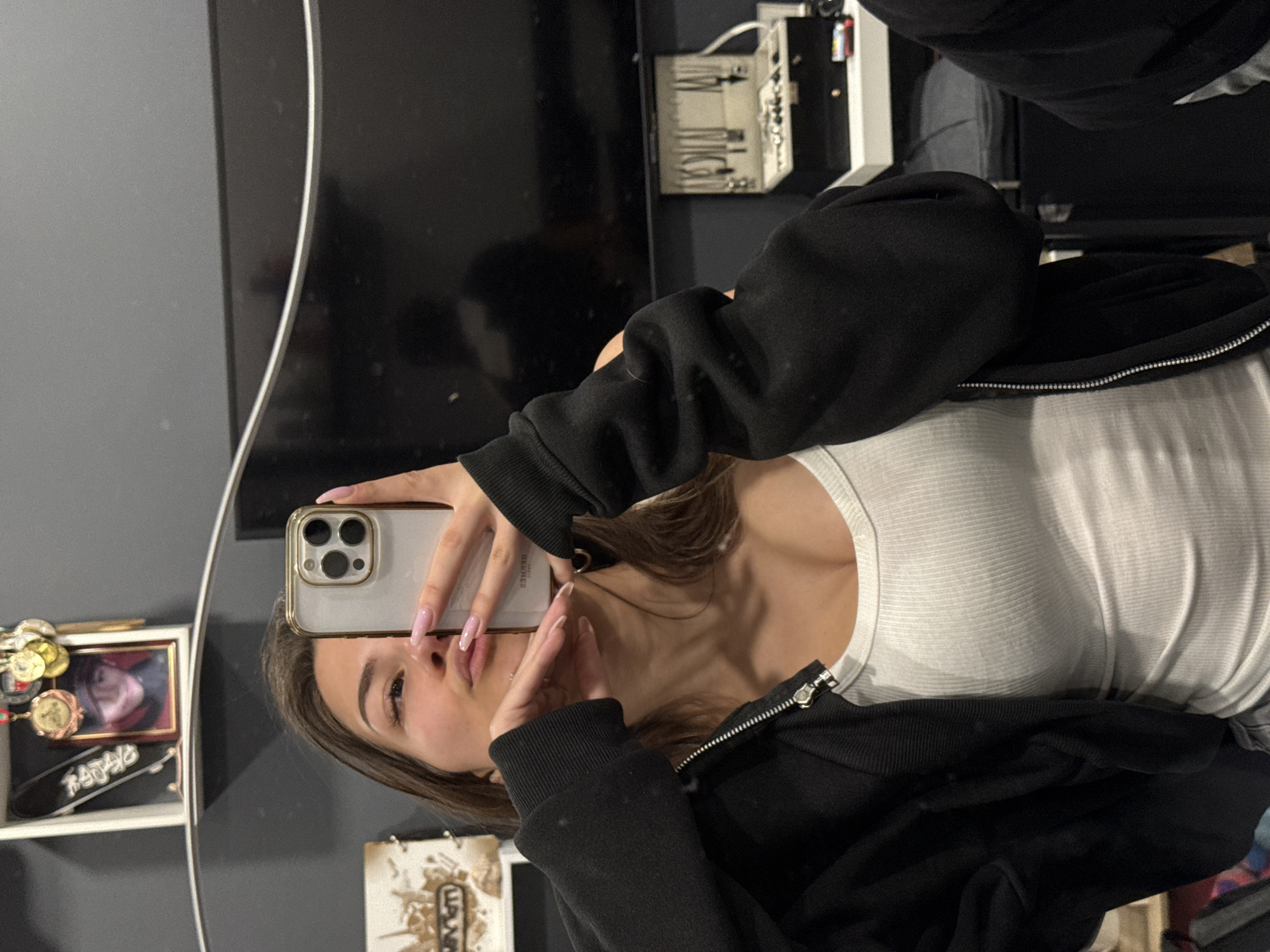 An image, rotated 90 degrees counter-clockwise, depicts a young woman taking a mirror selfie indoors in Sofia, Bulgaria. The woman, with medium-brown hair, is seen from the chest up, holding a light-colored smartphone with a triple-lens camera setup. She is wearing a white ribbed tank top and an open black zippered hoodie. Her nails are long, manicured, and painted a light pink or nude shade. She makes a "peace sign" or "V-sign" with her left hand, which is reflected in the mirror. Her expression is neutral, looking towards the phone.
The reflection in the mirror reveals elements of the room. To the left of her head, a white shelf unit holds several circular gold-colored medals or coins and a small framed photograph of a person. Below this, a black object with faint, reversed white text, possibly reading "RITUAL", is visible. In the background, behind her, a large dark flat-screen television or monitor is visible. Above it, electronic equipment with various ports and buttons is mounted. Part of a dark bed or couch can be seen in the lower reflected area. To the bottom right of the reflection, a piece of paper or card with ancient Egyptian hieroglyphs or similar symbols is displayed. The phone itself has "PULS" visible in reverse, and possibly "ULTRA" on its case. The setting appears to be a casual indoor space, likely a bedroom, with ambient artificial lighting.