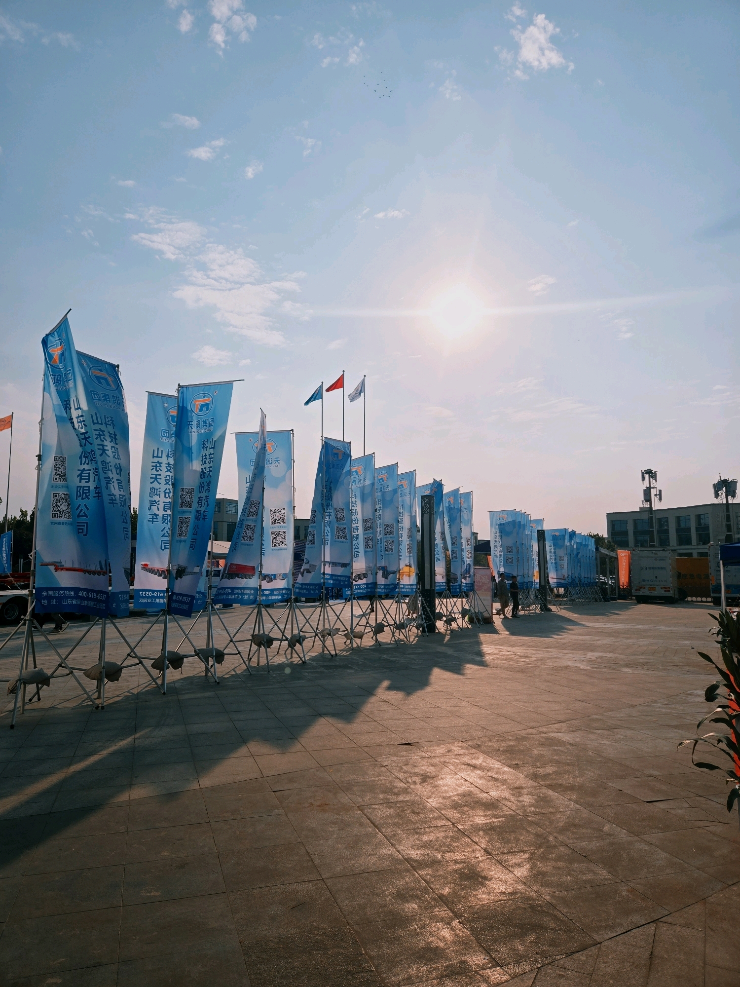 A line of blue and white flags bearing Chinese text and QR codes stands on metal tripods in an outdoor paved area. The flags are arranged in a row, creating a visual barrier. The sun is shining brightly in a pale blue sky with scattered clouds, casting long shadows from the flags onto the ground. In the background, there are buildings, cell towers, and a white truck. Two individuals are visible walking near the flags. The scene appears to be an outdoor exhibition or event, possibly in Liangshan County, China, given the presence of Chinese text on the flags and the overall aesthetic.