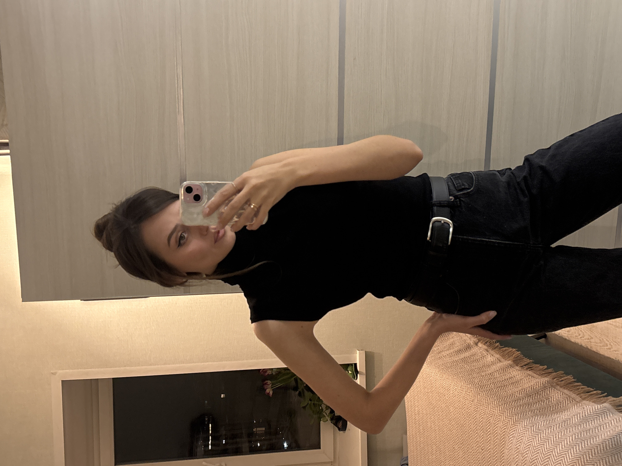 An adult female is depicted in a mirror selfie taken indoors within a residential apartment in Krasnoyarsk, Russia, during nighttime hours. The subject, positioned centrally, is wearing a black sleeveless turtleneck top, black jeans, and a black belt with a silver buckle. Her dark hair is styled in a bun, and a gold-colored ring is visible on her right ring finger. She holds a light-colored smartphone with a clear case in her right hand, extended towards the mirror for the selfie, while her left hand rests on her hip.
The immediate background on the left features a window revealing a dark exterior with blurred distant city lights. On the windowsill below, a vase containing purple flowers (likely tulips) and dark-colored footwear are present. To the right of the subject, and serving as the reflective surface, is a large, light-colored panel, possibly part of a wardrobe or sliding door, featuring a faint horizontal grey line. A portion of a bed, covered by a textured light brown or beige blanket with a herringbone pattern and fringed edge, is visible in the lower-left foreground. The bed frame appears to be dark green or teal. The room is artificially lit, contributing to a warm ambient light.