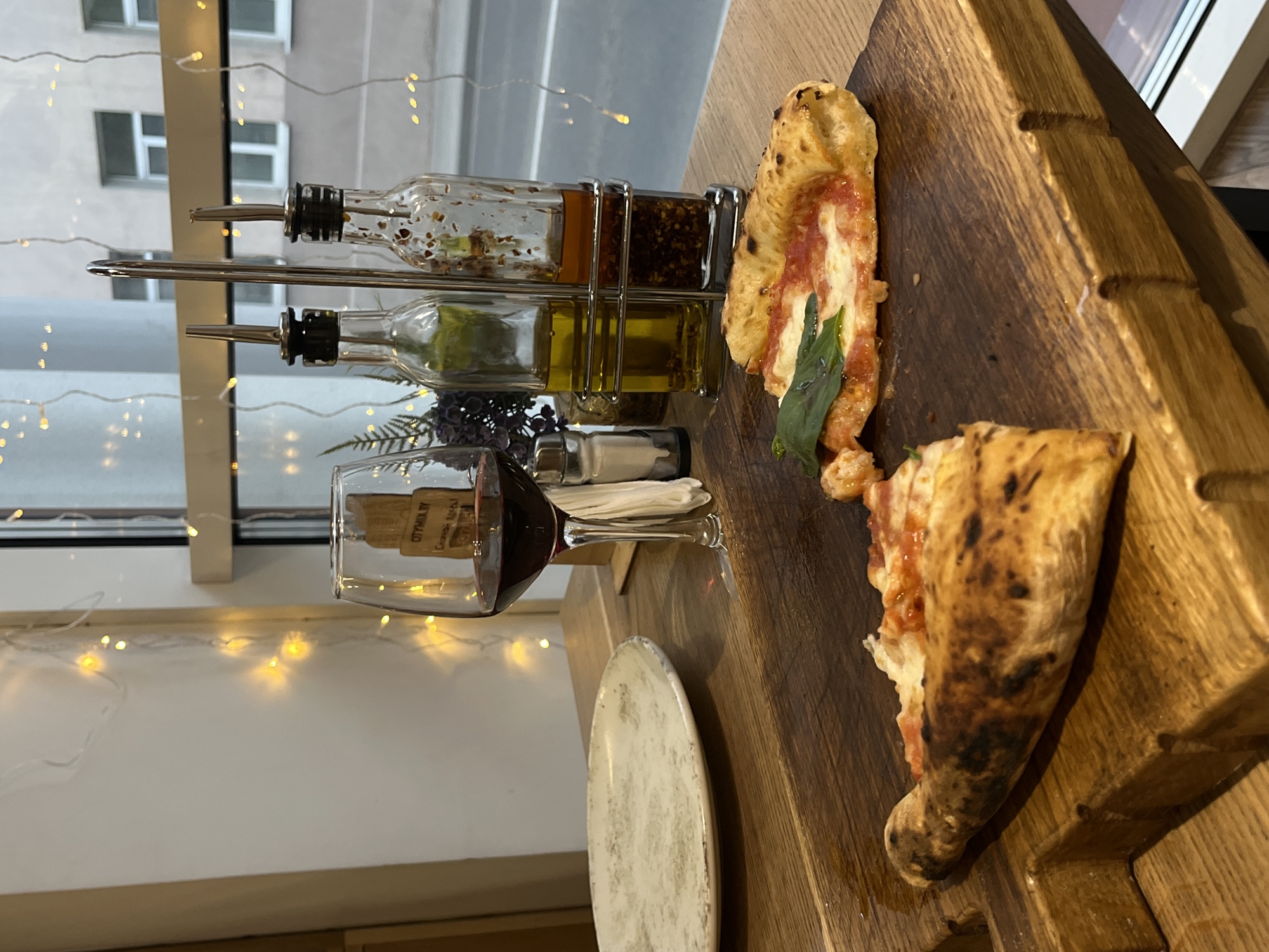 A casually staged still life captures a meal setting. A thick wooden cutting board displays two slices of pizza, one with a visible basil leaf, suggesting a Margherita style. Beside the pizza, a white plate and a glass of red wine are situated. In the background, a metal stand holds bottles of olive oil and spices. A window is visible, suggesting an interior location, with city buildings seen through the glass. Christmas lights are strung near the window, possibly indicating a festive or cozy atmosphere. The label on the wine glass displays “CITYMIX.BY” in Cyrillic, implying a location in Belarus, likely Hrodna.