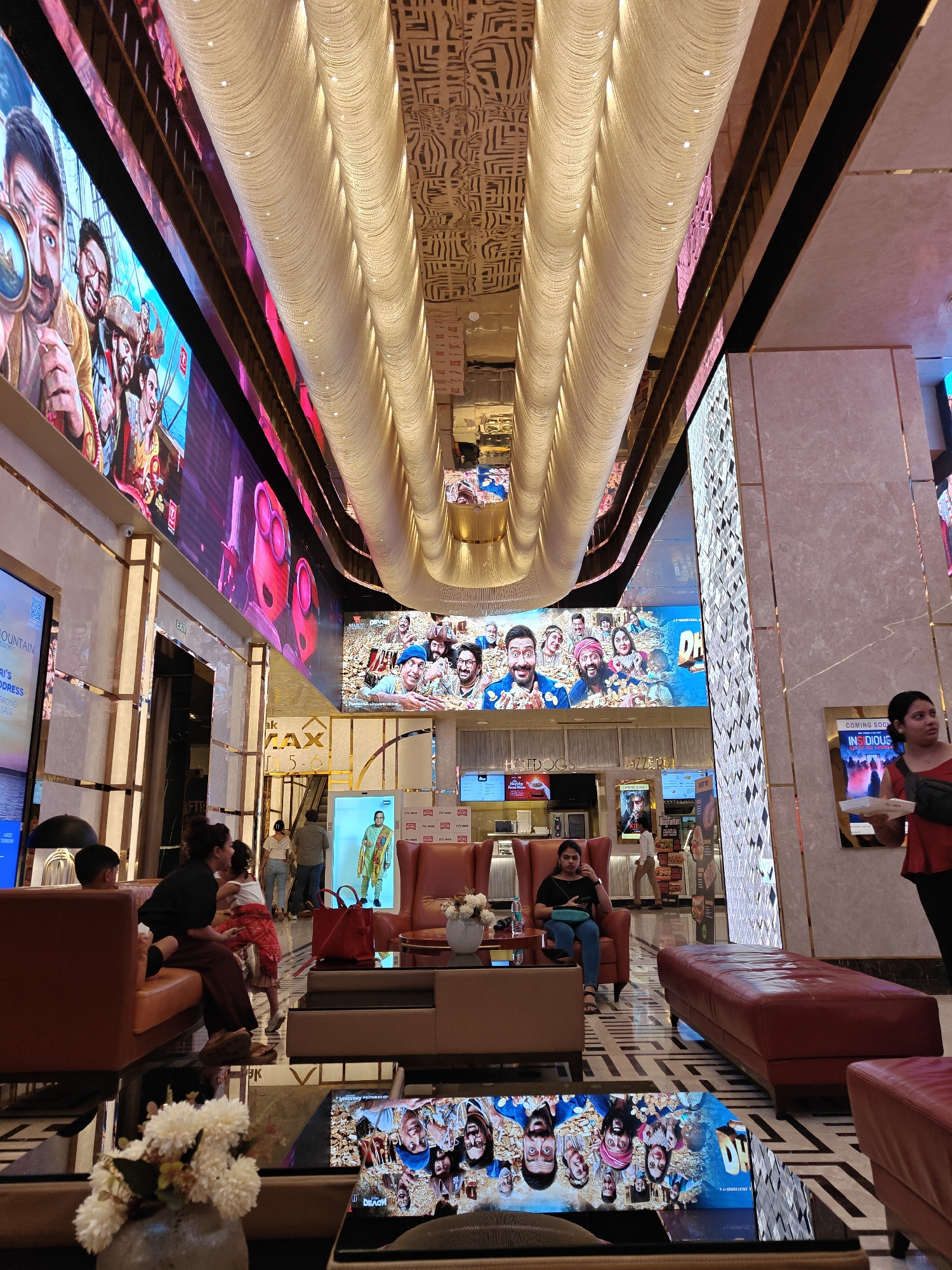The image captures the opulent interior of a multiplex cinema lobby in Mumbai, India. The scene is dominated by large LED screens displaying movie posters and trailers, showcasing vibrant imagery and text in Hindi. The ceiling features an intricate, illuminated design with a cascading effect, adding to the luxurious ambiance.
In the foreground, plush red leather sofas and armchairs are arranged for patrons. A coffee table in the center has a reflective surface, mirroring a movie poster of various faces. A vase with white flowers sits on a side table.
Several people are visible throughout the lobby. In the mid-ground, a group is seated on a sofa, including a woman in a black top and brown pants, and a child in a red outfit. A young woman in a black top and blue jeans is seated in an armchair, looking at her phone. Two individuals are standing near an escalator. In the background, more people are seen walking and interacting.
On the walls, large displays advertise movies, and signs indicate cinema services like "Hotdogs." The flooring is a glossy tiled pattern. The overall impression is a modern, well-lit, and sophisticated entertainment venue.