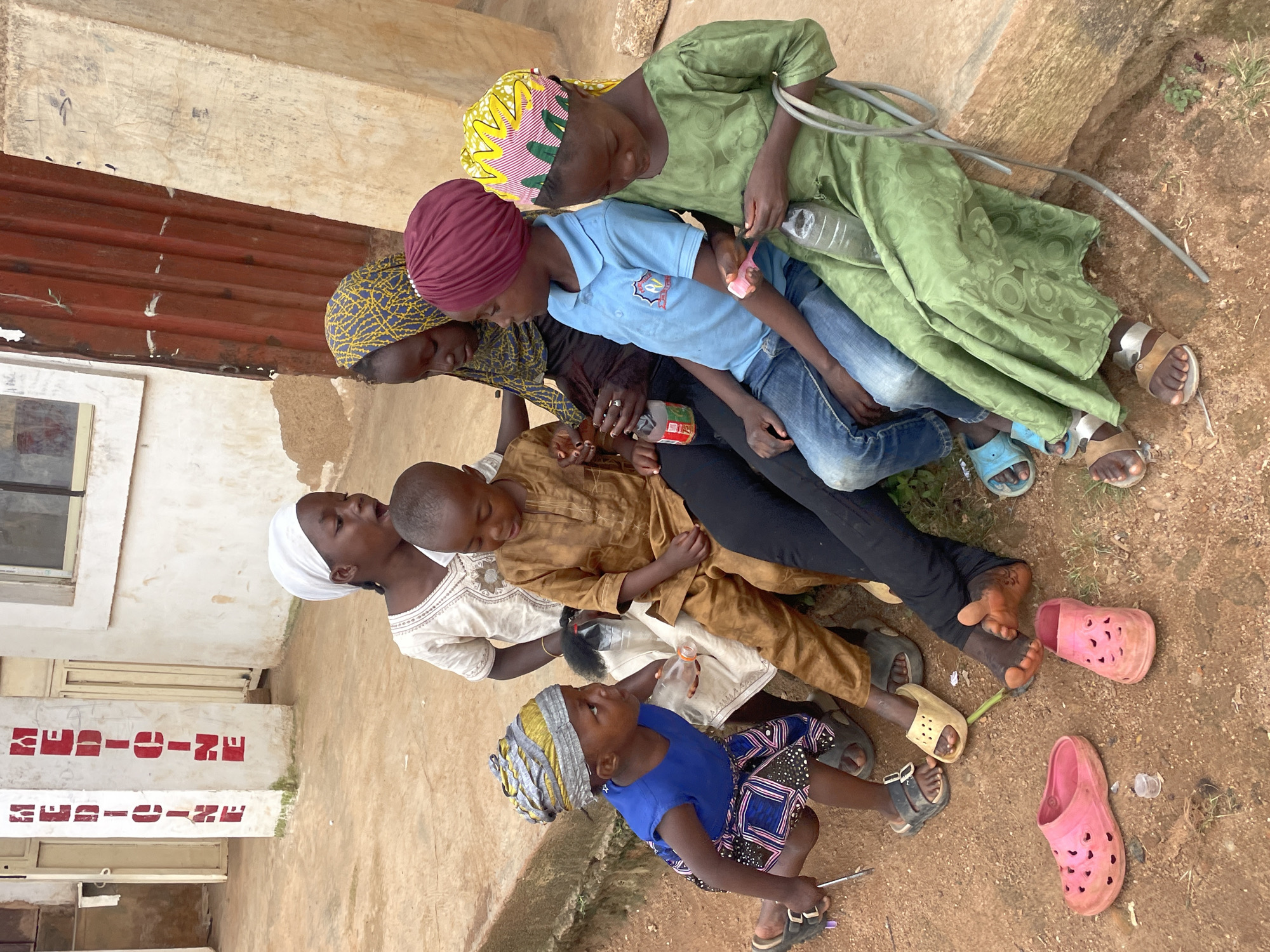 This photograph captures a group of six children sitting on a raised concrete ledge outdoors, likely in Jos, Nigeria, with a building that has the word "MEDICINE" stenciled in red on a pillar in the background. The children are of varying ages and are dressed in casual clothing.
In the foreground, a young girl in a blue sleeveless shirt and a patterned skirt is crouched down, holding a small object and looking to her right. Next to her, to the left, a young boy in a tan outfit is seated. Beside him, a slightly older girl in a white garment and white headscarf is also seated, looking to her left.
To the right of the young boy, a child wearing a blue collared shirt and blue jeans is seated, holding a clear plastic bottle with a white cap. Next to this child, a girl in a green patterned dress and a yellow and pink patterned headscarf is seated, holding a clear plastic bottle. In the background, partially visible behind the seated children, is another child wearing a blue and yellow patterned headscarf.
The ground is covered in reddish-brown dirt. Several pairs of colorful sandals and flip-flops are scattered around the children, including a pair of pink flip-flops in the lower left and a pair of yellow slides near the center. A thin metal rod or pipe lies on the ground near the edge of the ledge. The building in the background appears to be a simple structure with a window and a reddish corrugated metal wall. The overall scene suggests a casual gathering of children in an outdoor urban or semi-urban environment.