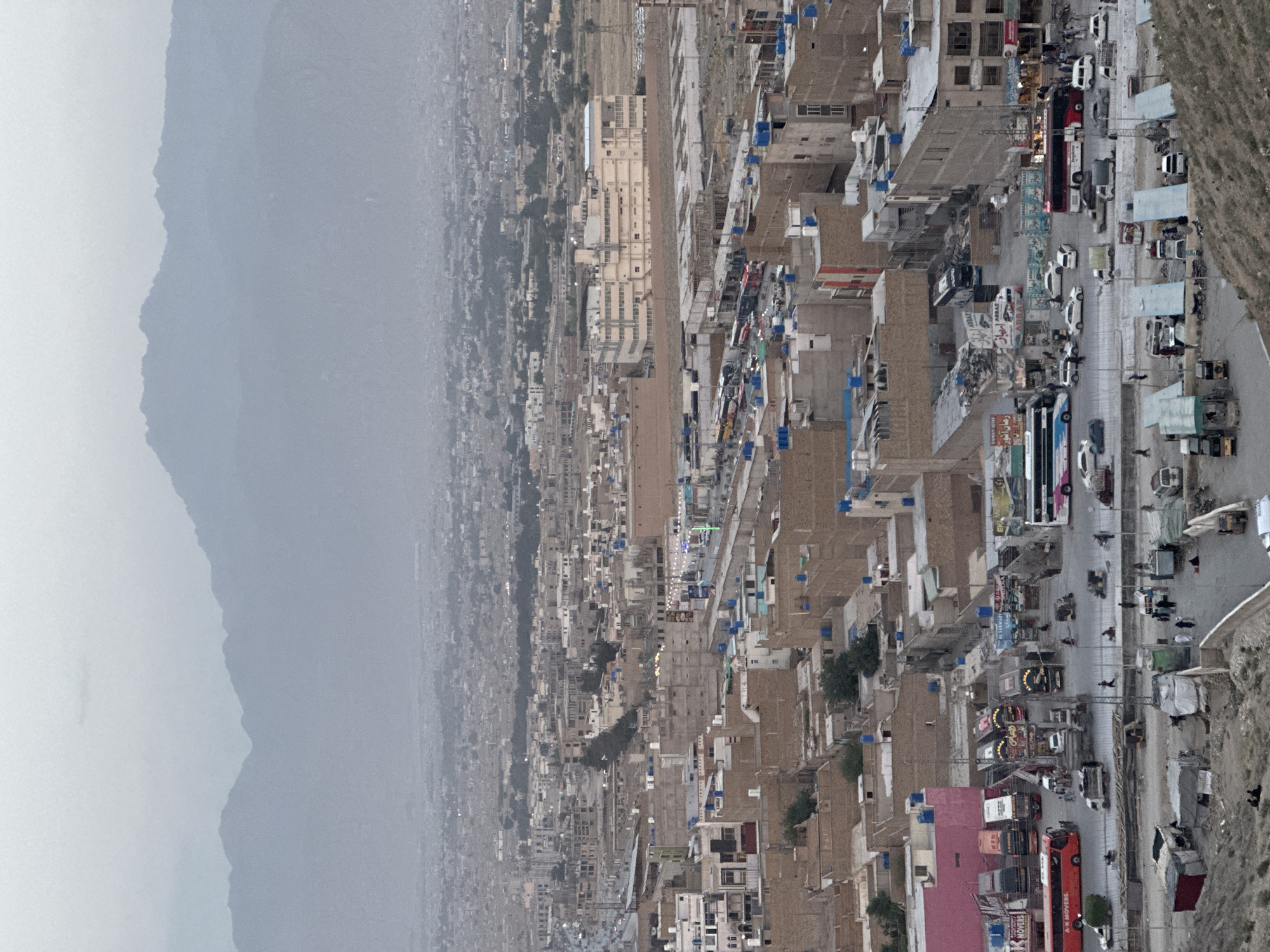 The image presents an aerial view of a densely populated urban area, likely within Quetta City Tehsil, Pakistan, given its architectural style and environmental context. Hazy mountains are visible in the left portion of the frame. Buildings are closely packed, primarily beige or light brown, with many displaying blue water tanks on rooftops. A main road lined with vehicular traffic, including buses, cars, and motorcycles, cuts through the cityscape. Signs and advertisements are visible alongside the road. The overall setting suggests a lower-income, high-density residential and commercial area within the city.
