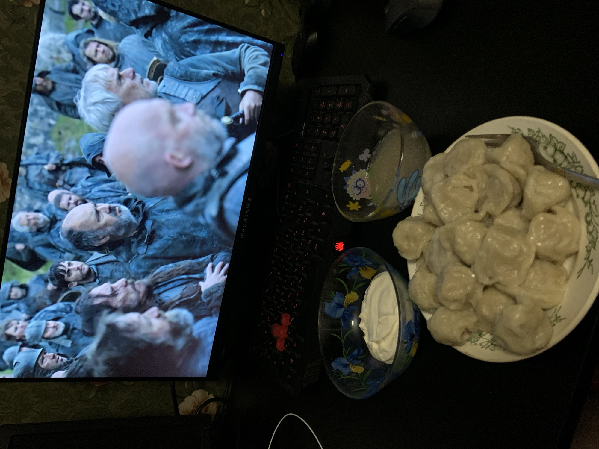 In the city of Desyatikhatka, Russia, an indoor scene captures a person in the foreground engaging in a meal while watching a screen. The meal consists of a plate of dumplings and two bowls, one containing a white creamy substance, possibly sour cream, and another with a light-colored liquid. A fork is placed on the plate of dumplings. Behind the meal, a computer monitor displays a scene from a media program, featuring a group of men in what appears to be a historical or fantasy setting, dressed in muted, dark clothing. The keyboard of the computer is visible, with some keys illuminated by red backlighting. The overall environment suggests a personal, indoor space.