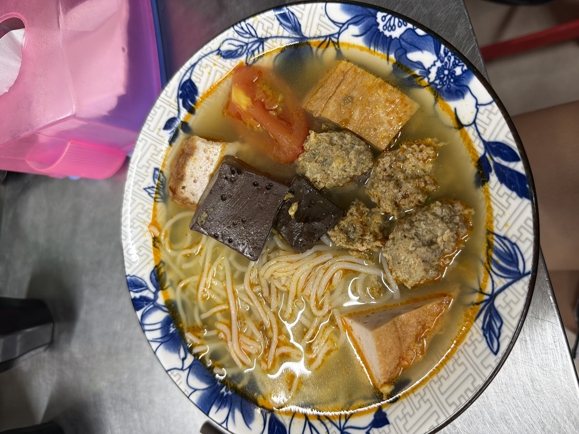 An overhead shot displays a bowl of Vietnamese noodle soup, placed on a grey, metallic table, likely stainless steel. The white ceramic bowl features a decorative blue floral pattern on its rim and a geometric, maze-like design accented with orange/gold on the inner edge.
The soup contains thin, white rice vermicelli noodles (bún) submerged in a clear, light yellowish broth, topped with flecks of orange oil or seasoning. Various ingredients are arranged in the bowl: several dark, rectangular pieces of congealed pig's blood, multiple irregularly shaped, greyish-brown meatballs (likely seasoned pork or fish paste), and light brown pieces resembling fried fish cake or tofu, some cut into rectangles and one into a triangle. Red tomato wedges provide a splash of color.
In the upper left of the frame, a translucent pink plastic container, possibly a tissue dispenser, is partially visible. The setting suggests a casual eatery or street food stall, consistent with the given location context of Đà Nẵng, Vietnam. No people are visible, and there is no discernible text in the image.